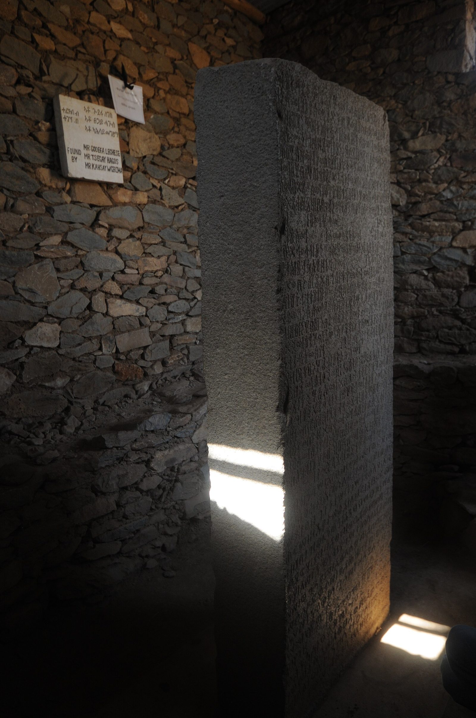 A tall grey granite slab standing upright in a dim room with rough stone walls, one face densely covered in carved inscription running its full height. A slot of daylight falls across the floor beside it. On the wall behind hangs a small hand-lettered placard carrying script in one alphabet and, in English capitals, the words FOUND BY followed by three men's names.