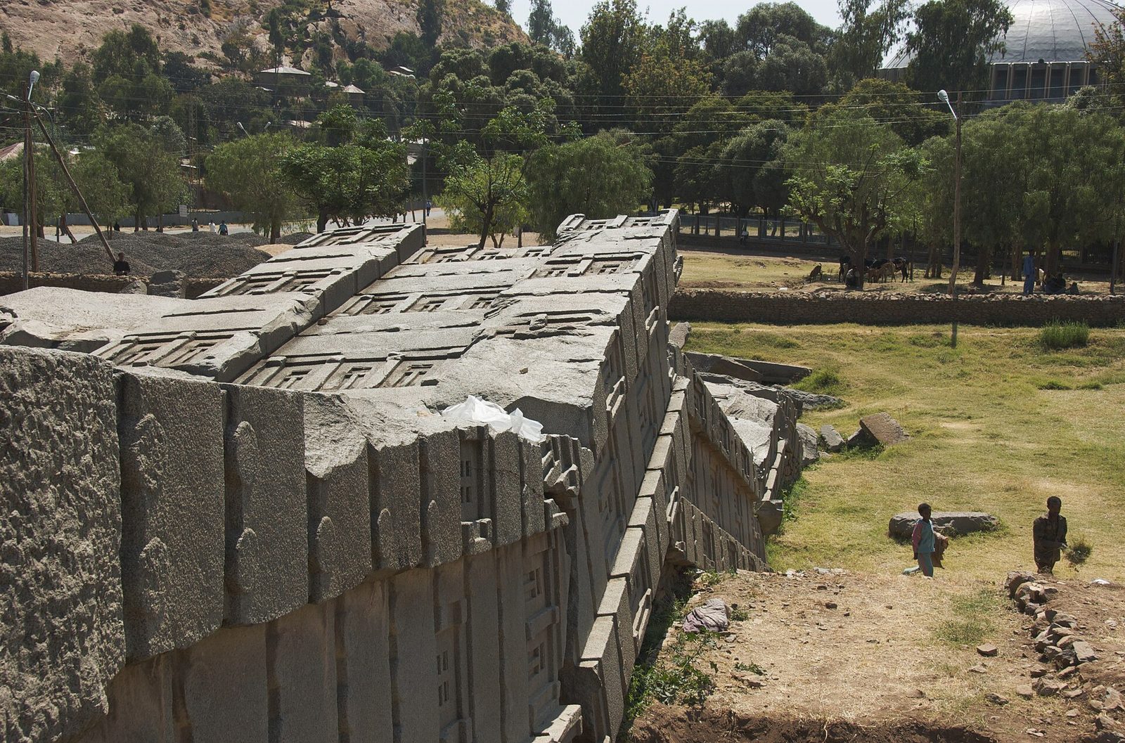 A colossal granite monolith lying broken on the ground in massive sections, its upper face carved with tier upon tier of false windows and projecting beam-ends imitating a multi-storey building. Two children stand on the grass at the right, dwarfed by it. Behind are trees, power lines, low buildings, grazing animals, and the dome of a modern church at the upper right.