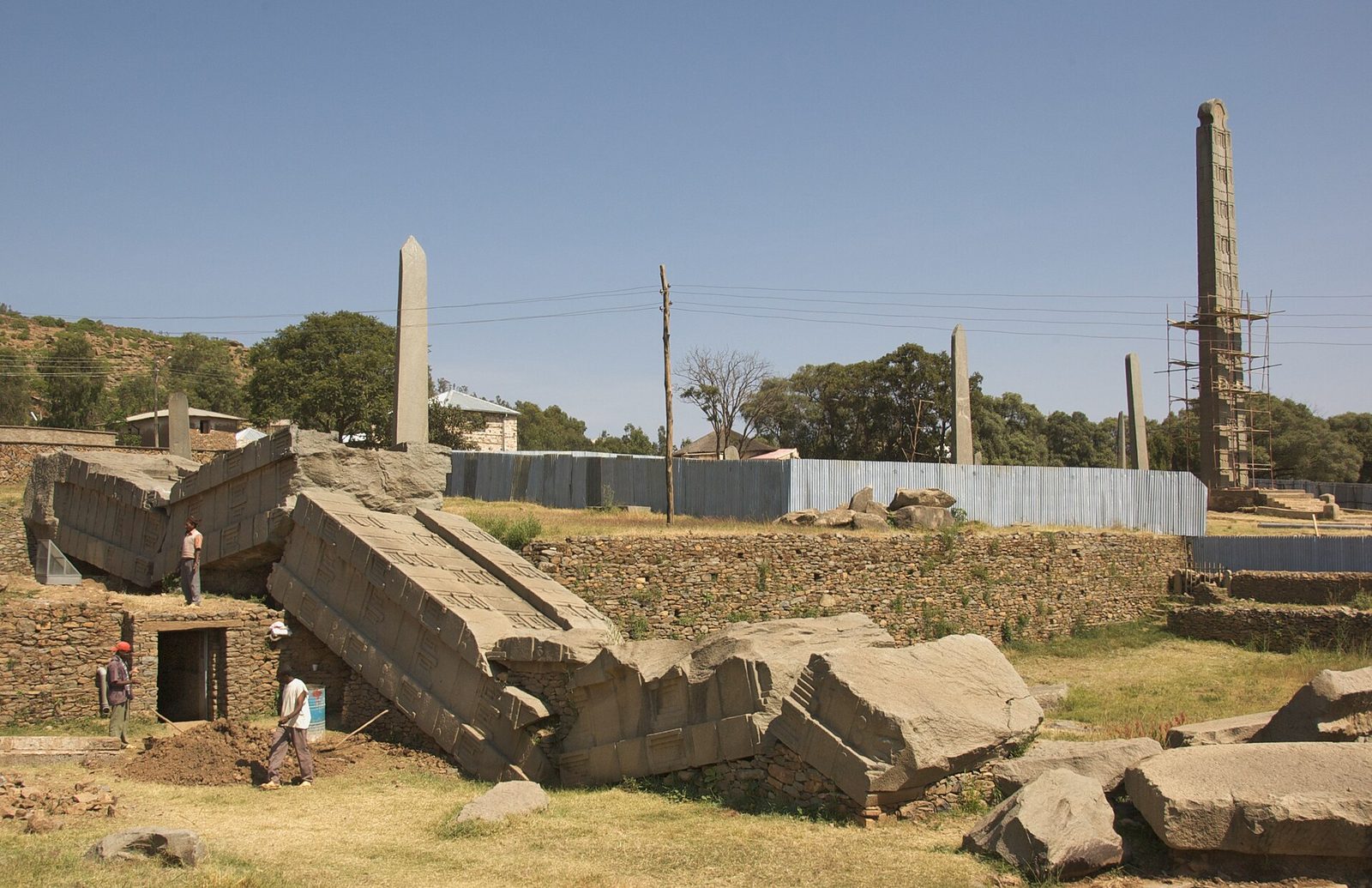 A field of stone monuments. A colossal carved monolith lies broken across the foreground in massive sections. Several plain, uncarved stelae stand upright among them. At the right a stela is encased in scaffolding. Corrugated-iron hoarding runs across the middle distance, with terraced dry-stone walling, a dark doorway into an underground chamber at the left, and workers moving among the stones.