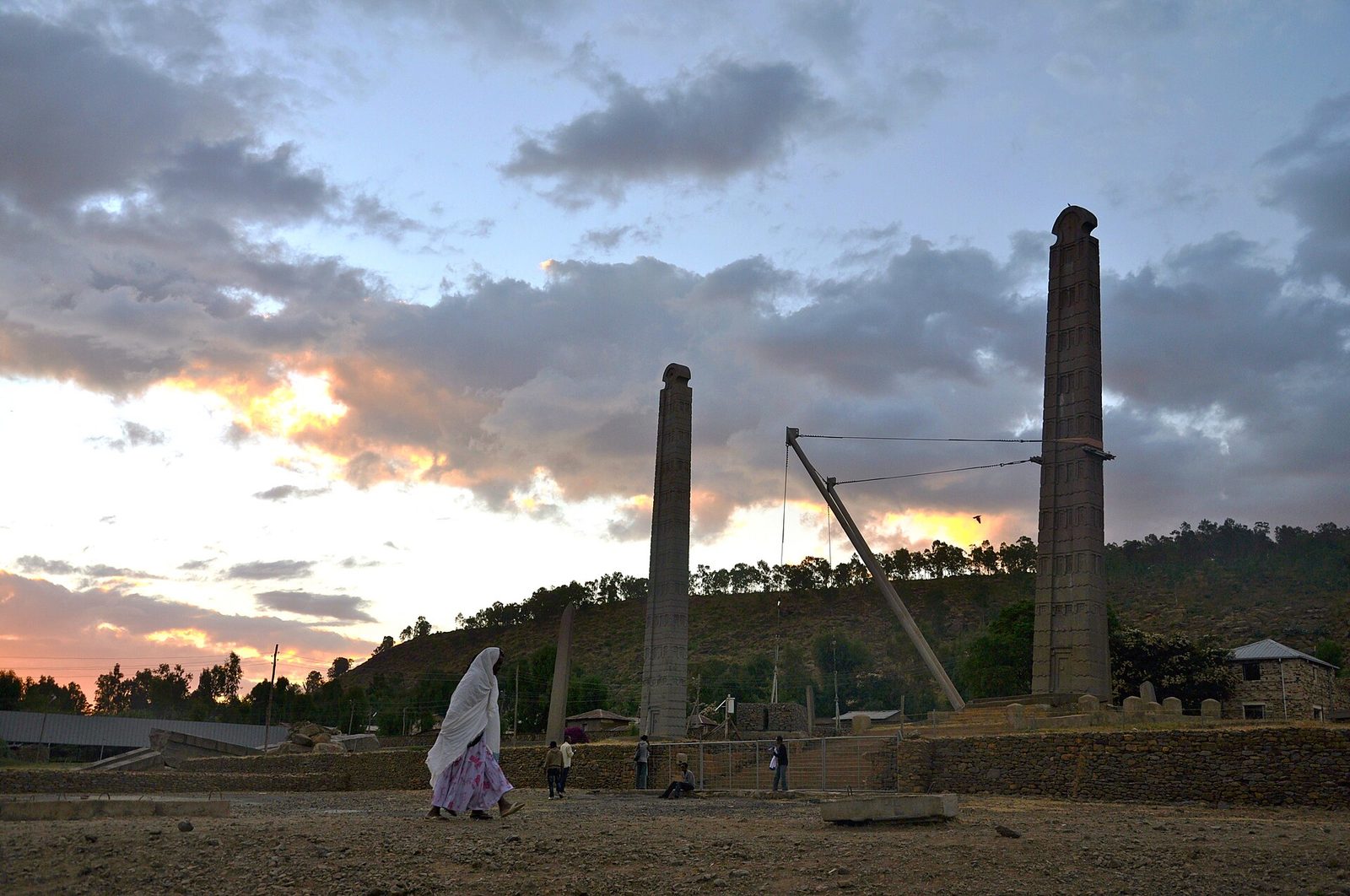 Two tall carved stone monoliths standing against a sunset sky of broken cloud, each cut with tiers of false storeys and finished with a rounded arched head. The taller one at the right is braced by a steel A-frame and guy cables. A woman in a white shawl walks across the foreground and several people stand near the bases. Low stone walling, a fence and wooded hills lie behind.