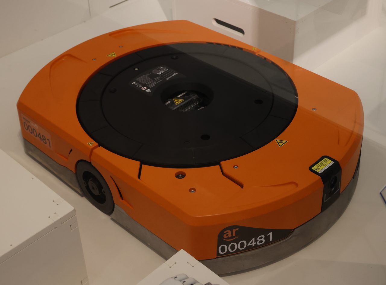 A low, flat, wheeled warehouse robot photographed on a pale panelled surface behind a glass edge, in what reads as a display case rather than a working aisle