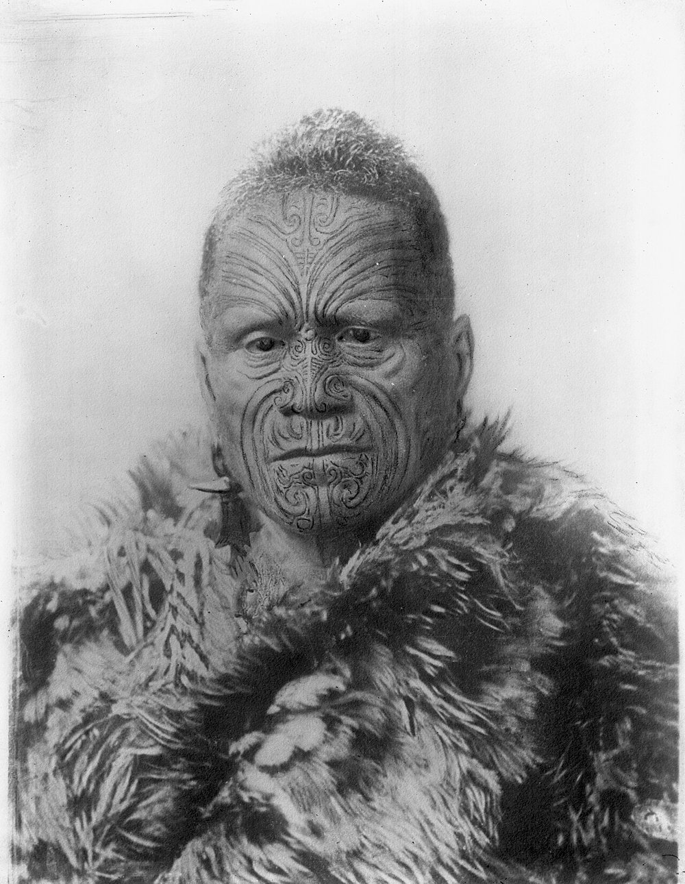 A black-and-white photograph from around 1894 of a Maori man wearing a feather cloak, his face covered with intricate spiral and curved ta moko tattoo patterns across the forehead, cheeks, nose, and chin