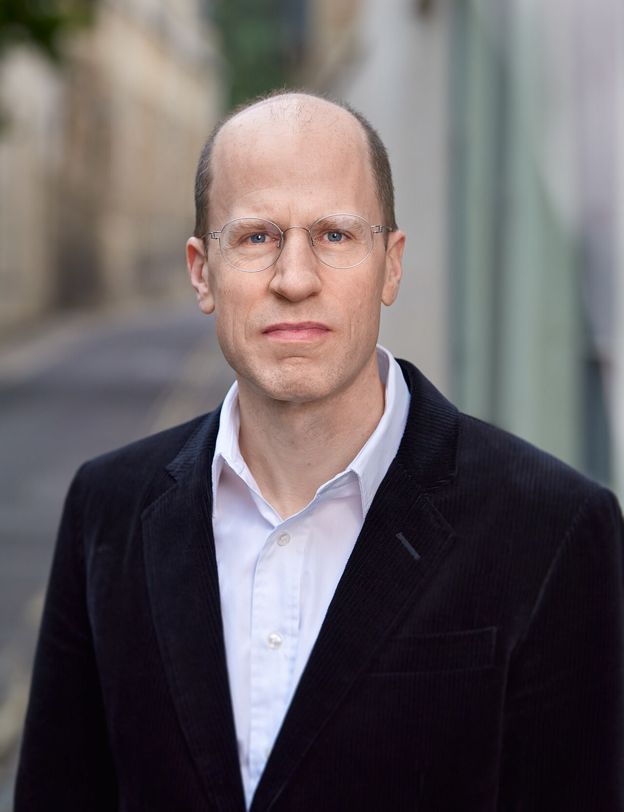A portrait photograph of the philosopher Nick Bostrom, balding, in round wire glasses and a dark corduroy jacket over an open-collared white shirt, against a blurred street background
