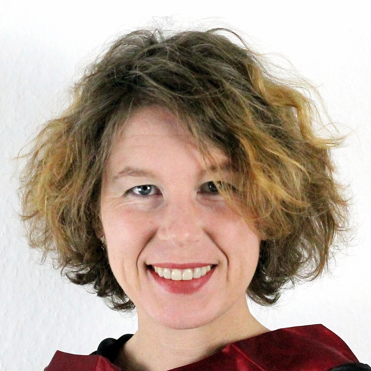 A portrait photograph of the physicist Sabine Hossenfelder, with wavy shoulder-length hair, smiling, in a dark red top against a white background