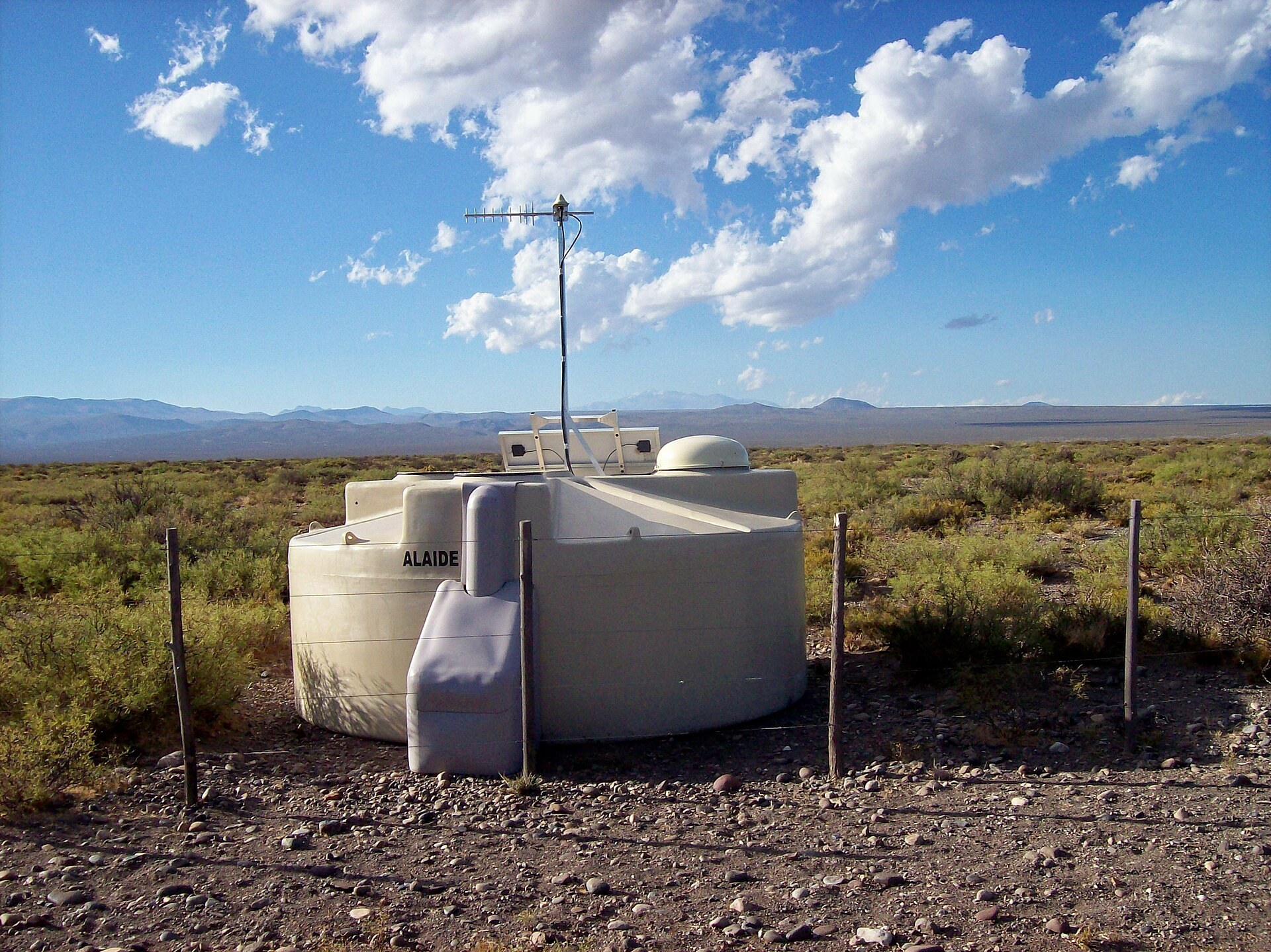 A white cylindrical cosmic-ray detector tank with a solar panel and antenna on top, standing in scrubby grassland with distant mountains and a blue cloudy sky, at the Pierre Auger Observatory in Argentina
