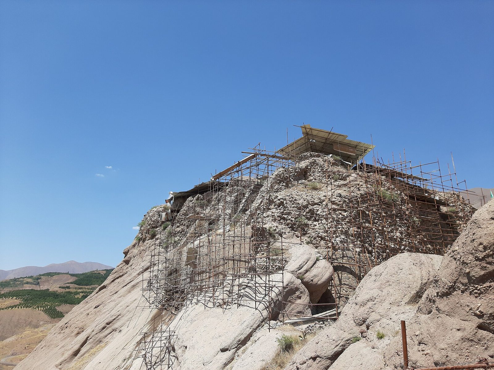 A pale grey-white rock ridge under a clear blue sky, its whole upper flank covered in a dense lattice of rusting steel reinforcing bars forming a conservation scaffold. A modern corrugated shelter roof on posts stands on the summit. Very little masonry is visible: a few courses of stone and a small arched opening low at the right. A green cultivated valley and brown hills lie away to the left.