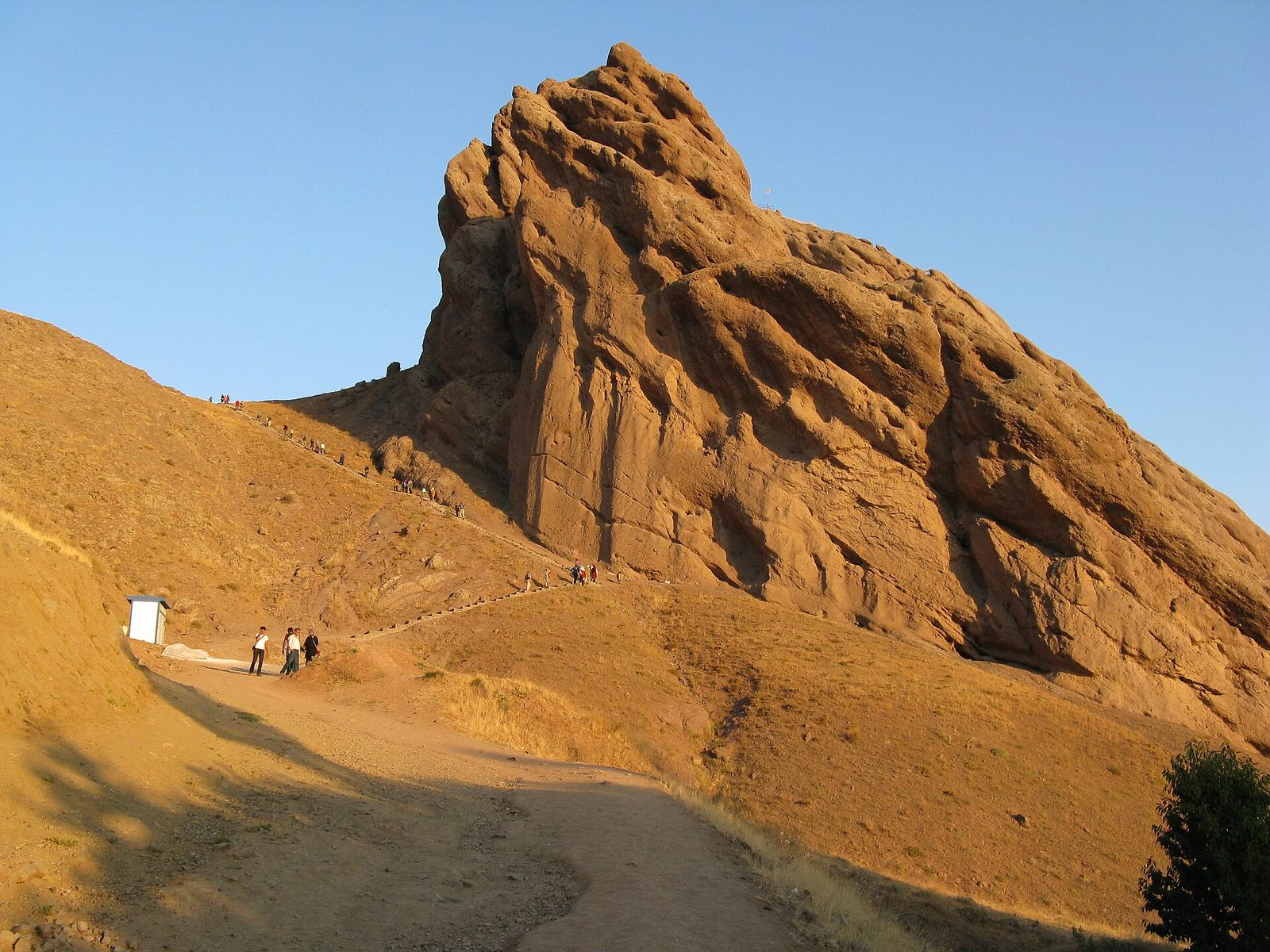 A huge tawny sandstone crag rising from dry grassy slopes against a clear blue sky in low golden light. A dirt track runs up from the foreground and a stepped path with a handrail is traced along the flank, with about twenty small figures strung out along it and along the ridge above. A small white kiosk stands at the left with three or four people beside it, and a dark green tree at the lower right. A tiny flag flies on the summit.