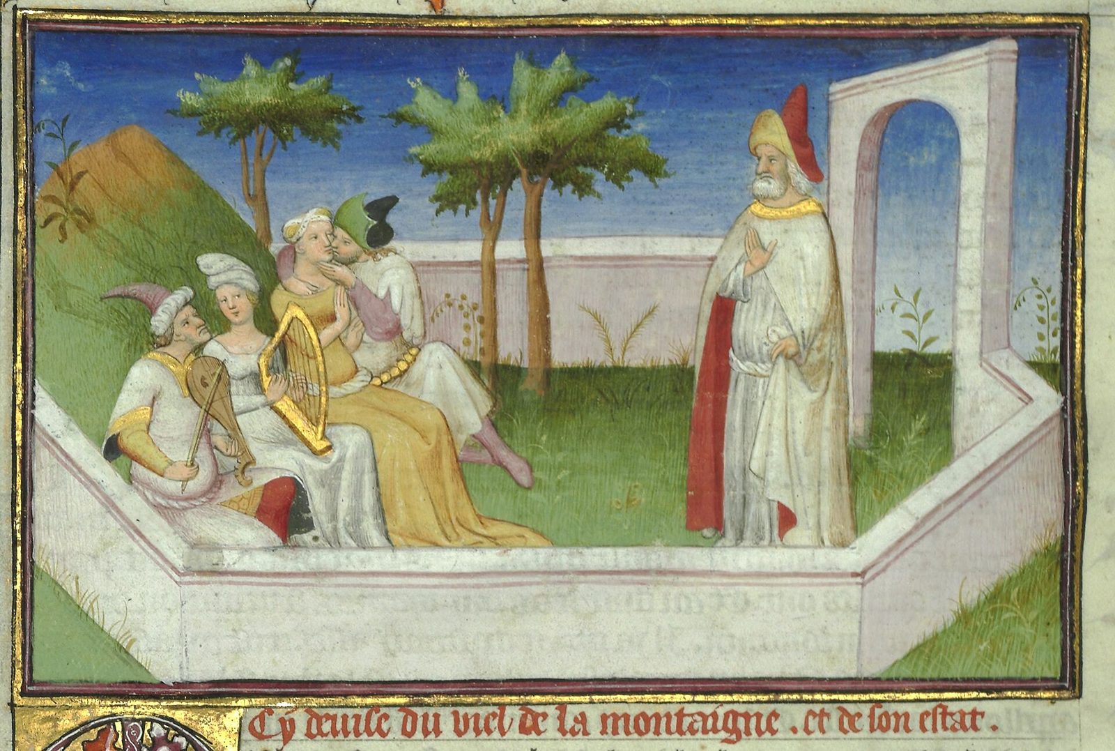 A French illuminated manuscript miniature in a gold and red frame. Inside a low white and pink walled enclosure on green grass sit four young people in Western European court dress: a man in a red and white hood playing a lute with a plectrum, a woman in white, a woman in a gold gown holding a gold harp, and a couple embracing and kissing. Two stylised trees stand against a deep blue sky. At the right, outside the parapet beside a white arched gateway, an old bearded man in white robes with a red-lined cloak and a yellow and red turban raises one hand. Beneath the frame runs a line of red medieval French naming the Old Man of the Mountain.
