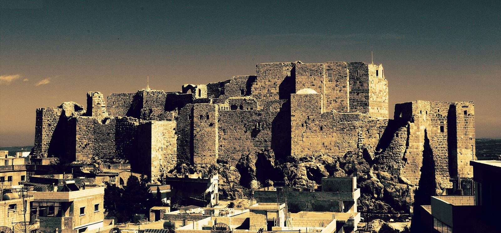 A large stone fortress standing substantially intact on a rocky outcrop, with curtain walls, square towers, a domed structure and sections open to the sky at either end. It rises directly above a modern town of flat-roofed concrete houses with rooftop water tanks and satellite dishes, with a cypress and other trees in between. The photograph is strongly toned in sepia and teal with a darkened sky.