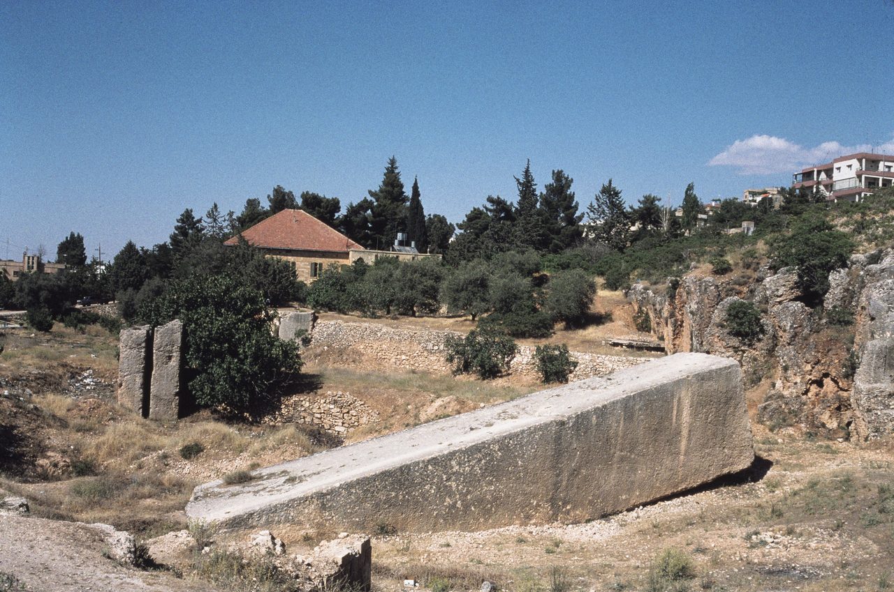 A colossal rectangular limestone monolith lying at a shallow angle in the Baalbek quarry, photographed in 1996, with the cut quarry face at right and the town of Baalbek on the ridge behind
