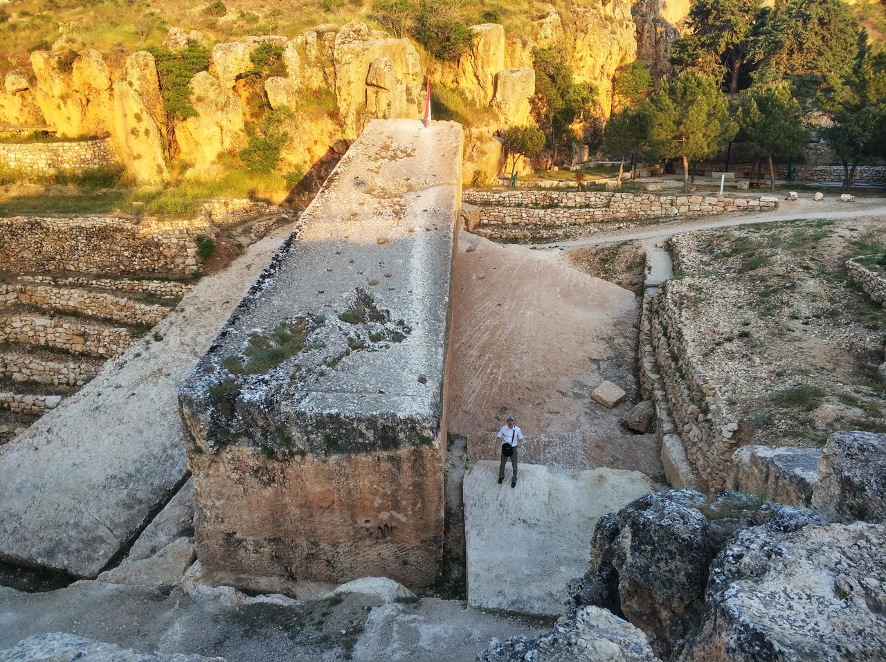 An elevated view of a colossal partially quarried limestone monolith lying in the Hajjar al-Hibla quarry at Baalbek, with a man standing at its lower end for scale