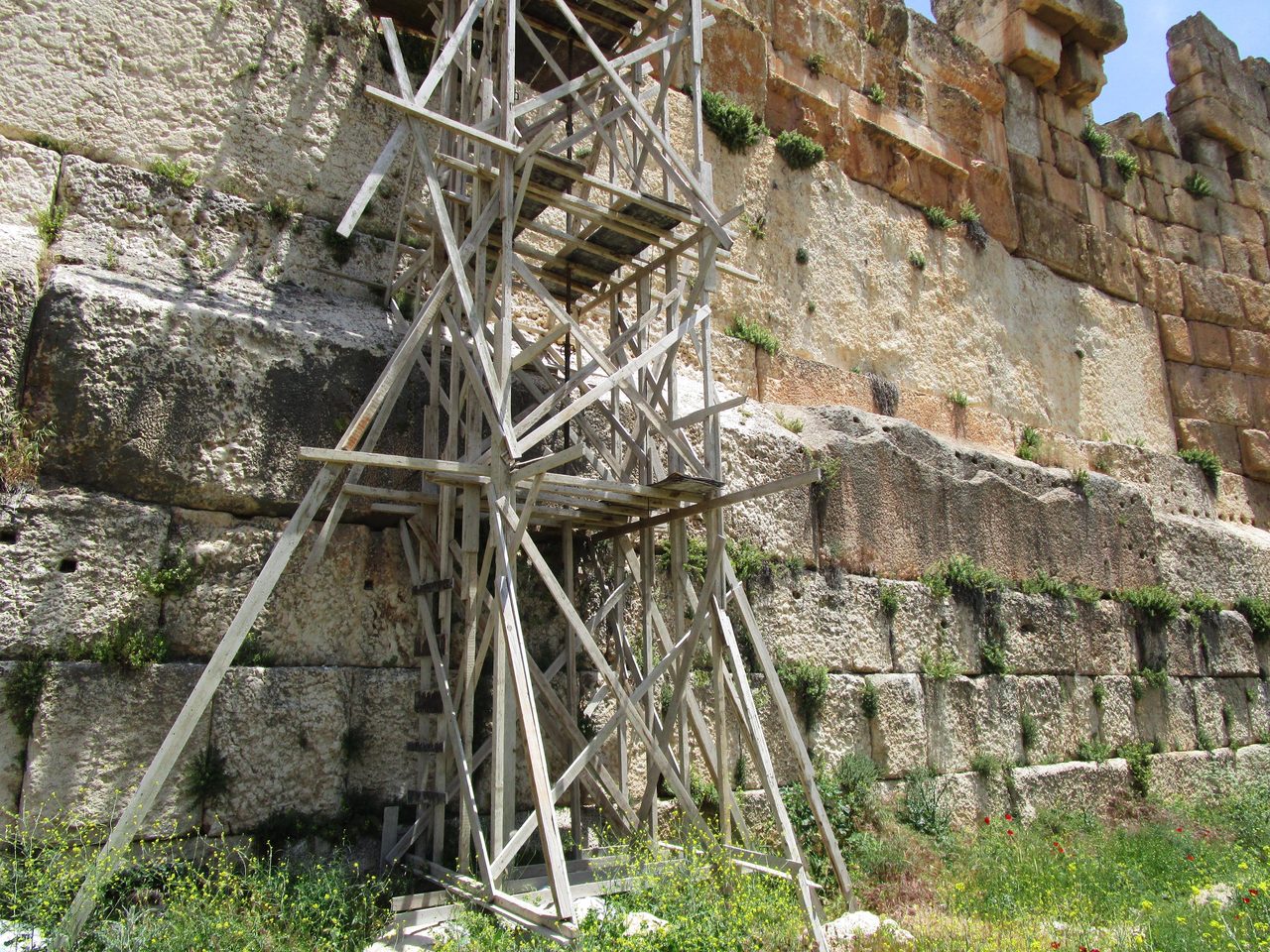The western podium wall of the Temple of Jupiter at Baalbek seen from ground level, with a tall modern timber scaffold tower standing against it