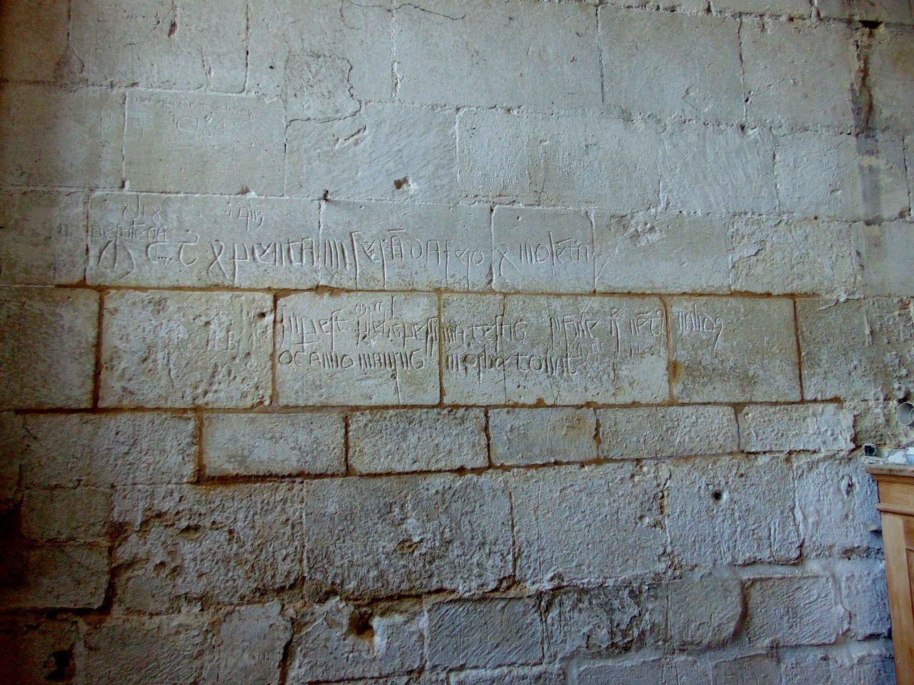 A wall of pale ashlar blocks inside a church tower, with several lines of shallow medieval letters scratched across two courses in a cramped angular hand, much of it worn and broken by the joints between the blocks