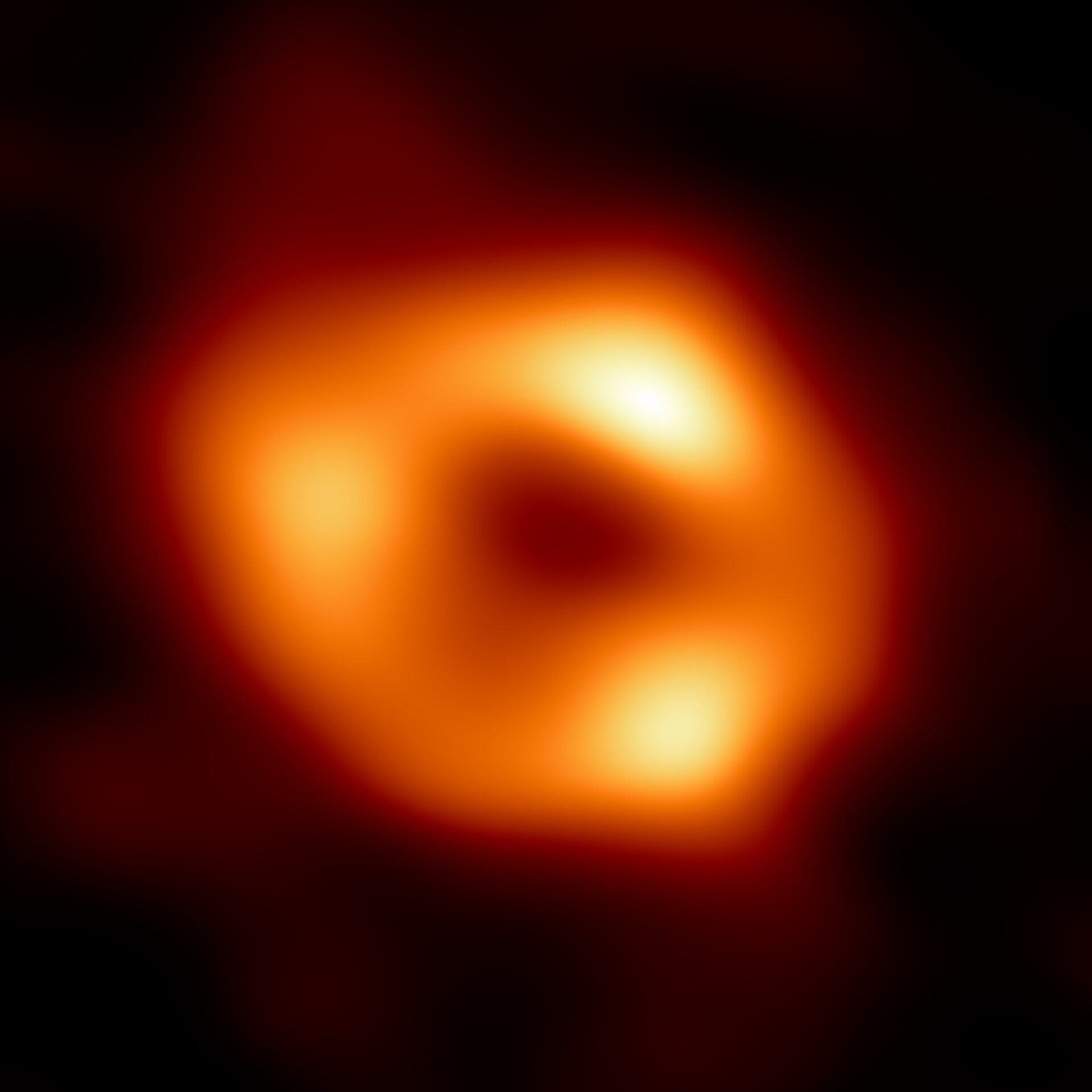 A radio-telescope image of a glowing orange ring with several brighter knots around a dark central region, against black, softer and less sharply defined than the M87 image