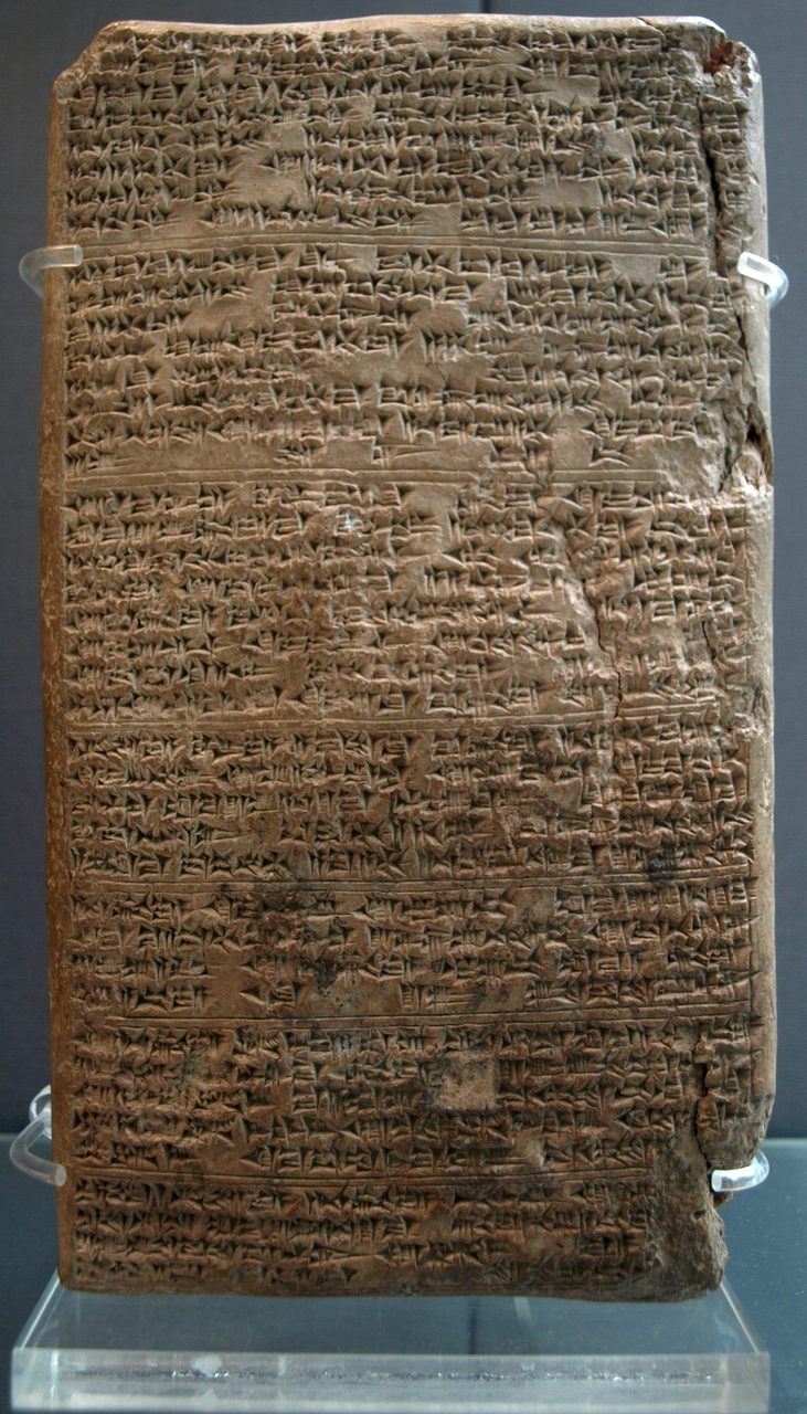 A tall rectangular clay tablet the colour of red brown earth, its whole face covered in dense horizontal rows of small cuneiform wedges separated by a few ruled dividing lines, chipped and cracked down the right edge, standing upright in clear plastic clips on a clear mount against a dark background