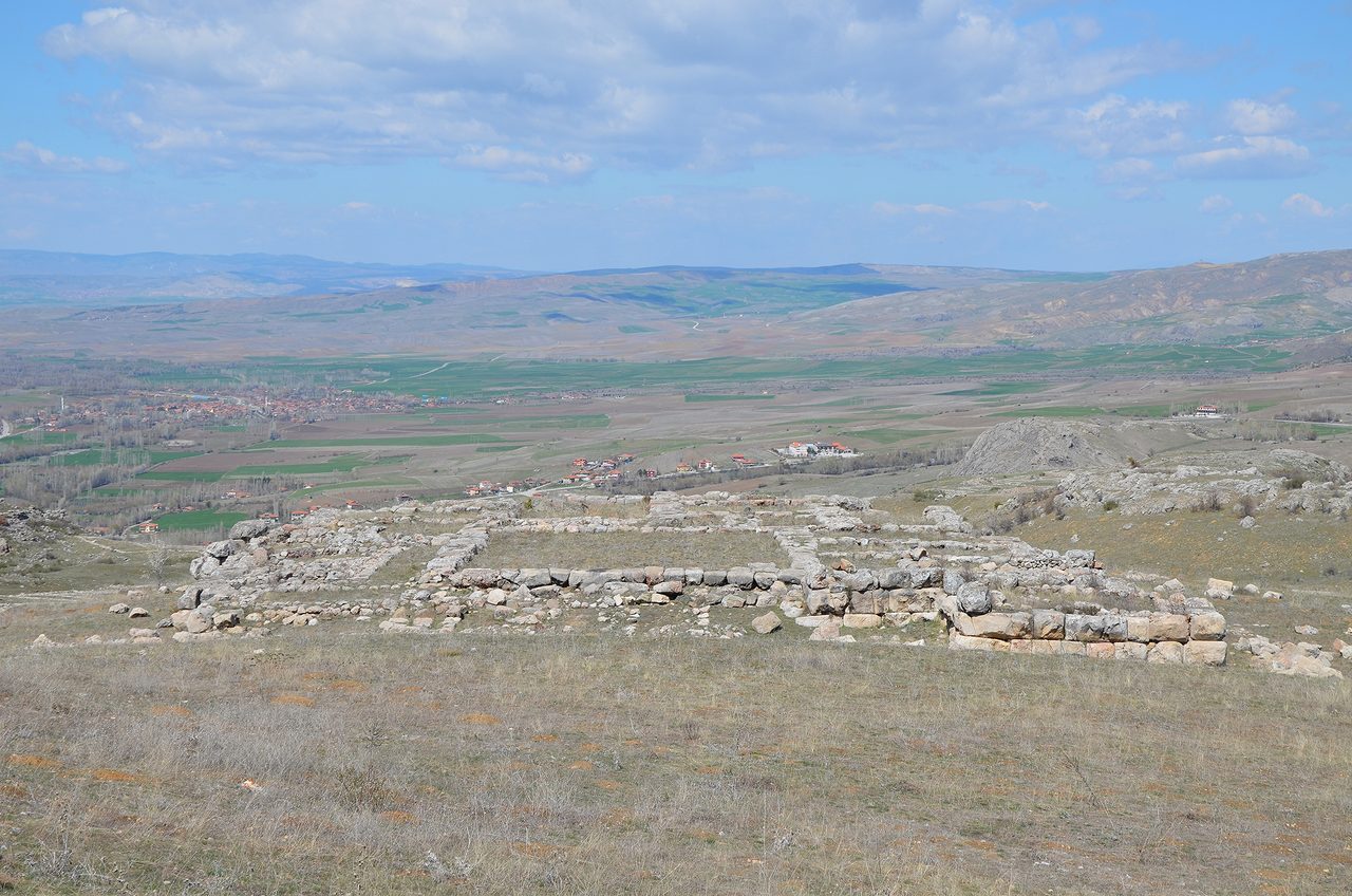 A hillside of dry grass carrying low stone foundation walls that outline rectangular rooms barely above the turf, with a broad valley beyond holding green and ploughed fields, a modern village of red roofed houses in the middle distance, and low hills under a blue sky