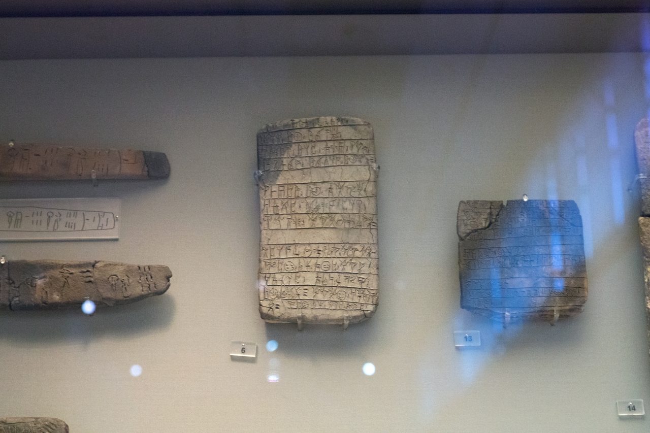 A museum case photographed through glass, with reflections and small bright spots across it, holding several clay tablets mounted on a pale grey wall: two narrow strip shaped tablets at the left with a white panel between them carrying an ink line drawing of a tablet and its signs, a large page shaped tablet at the centre ruled into horizontal lines of incised signs with its top left corner discoloured dark, and a squarer chipped fragment at the right, with small numbered cards below them