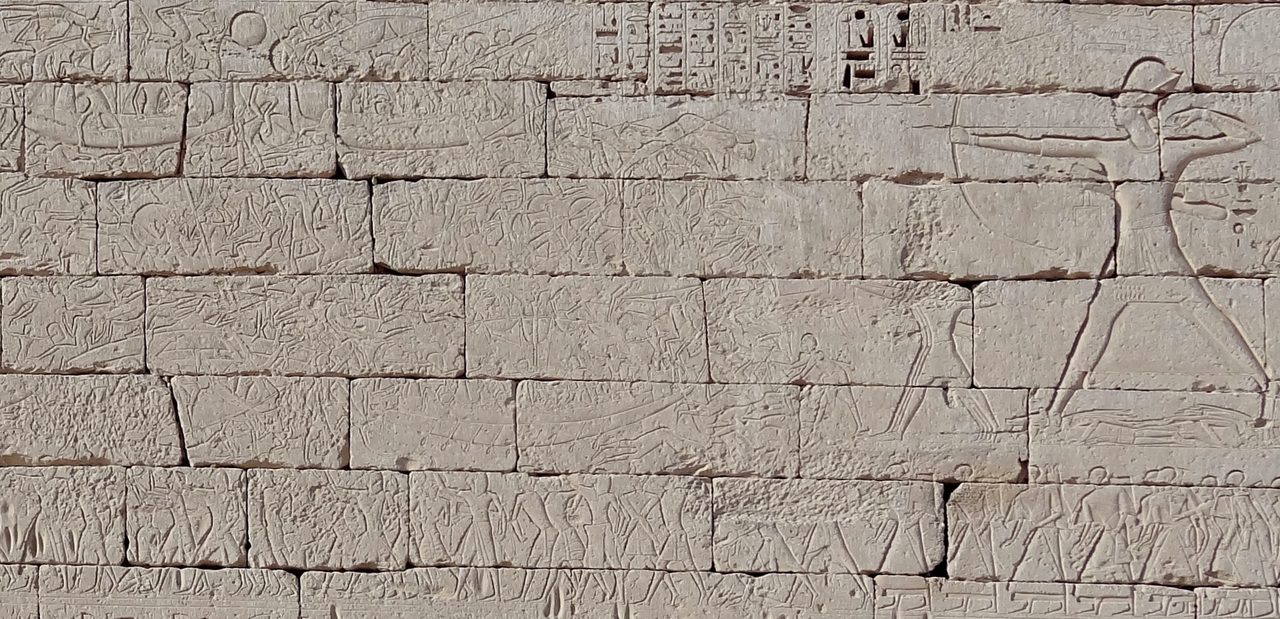 A wide expanse of pale limestone wall in coursed blocks, carved in shallow worn relief: at the right a single figure many times the size of the rest strides forward drawing a bow at full stretch, while across the rest of the wall dozens of small figures are packed together, many of them sprawled or upside down, with a few long curved hulls among them and a row of small upright figures along the lowest band