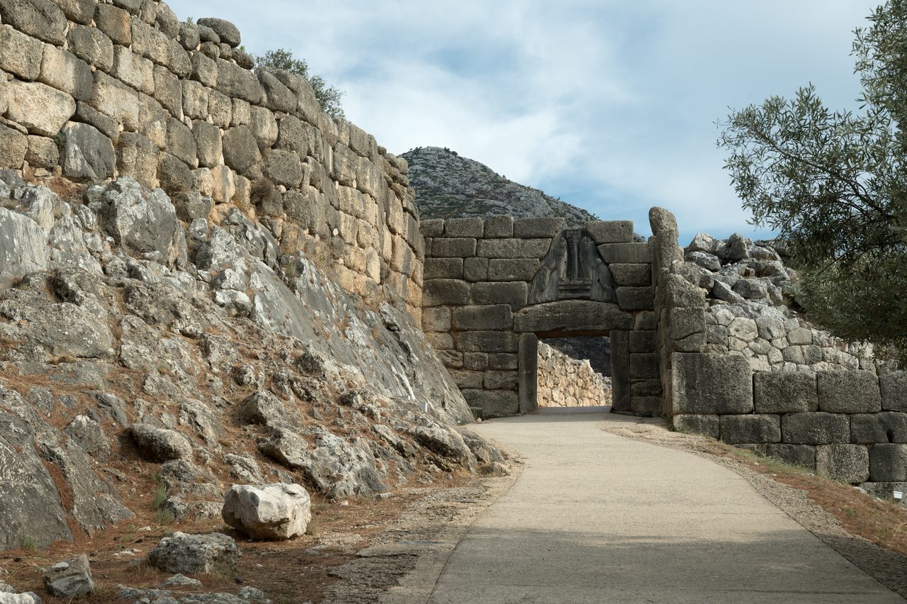 The approach to a monumental stone gateway built from huge fitted blocks, with a triangular carved slab above the lintel showing two animals standing on their hind legs facing each other, forepaws on a stepped base and a column between them, both animals ending at the shoulders with no heads, and a smooth modern path running up to and through the opening