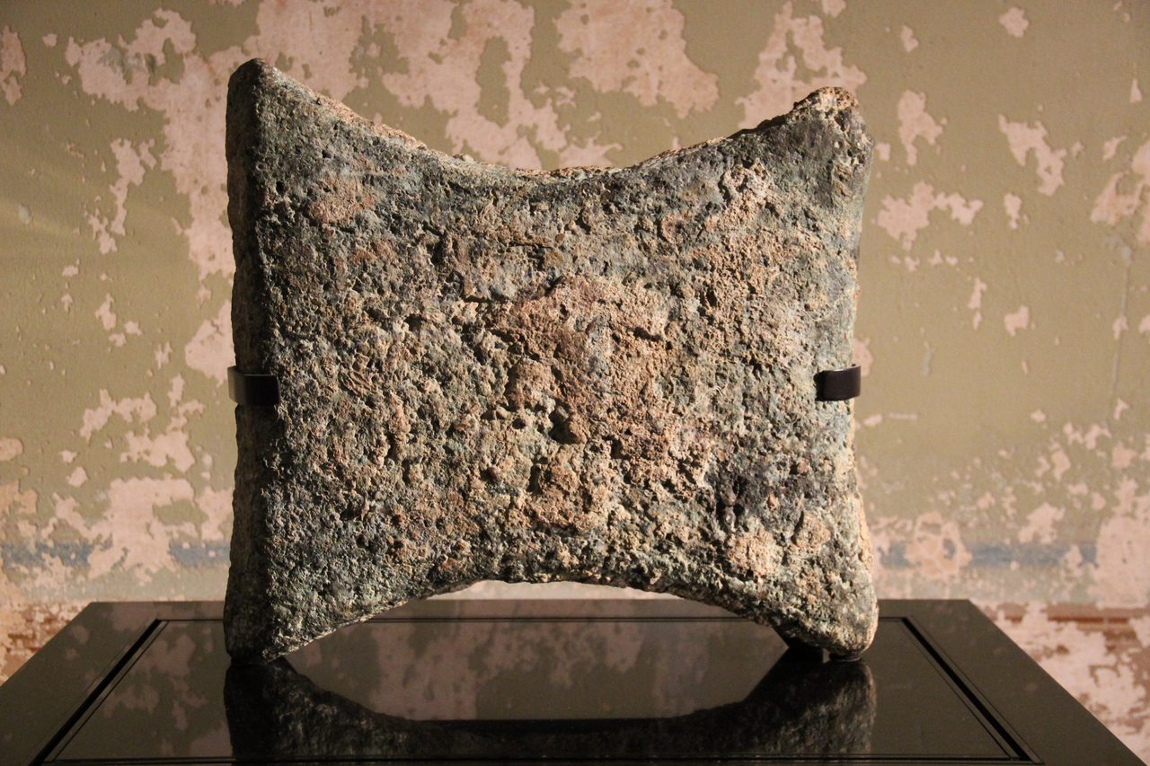 A roughly square metal slab with four projecting corner handles and concave sides, standing upright on a dark polished plinth and held by two small clips, its surface corroded to a mottled green and rust orange, against a wall of peeling mottled plaster