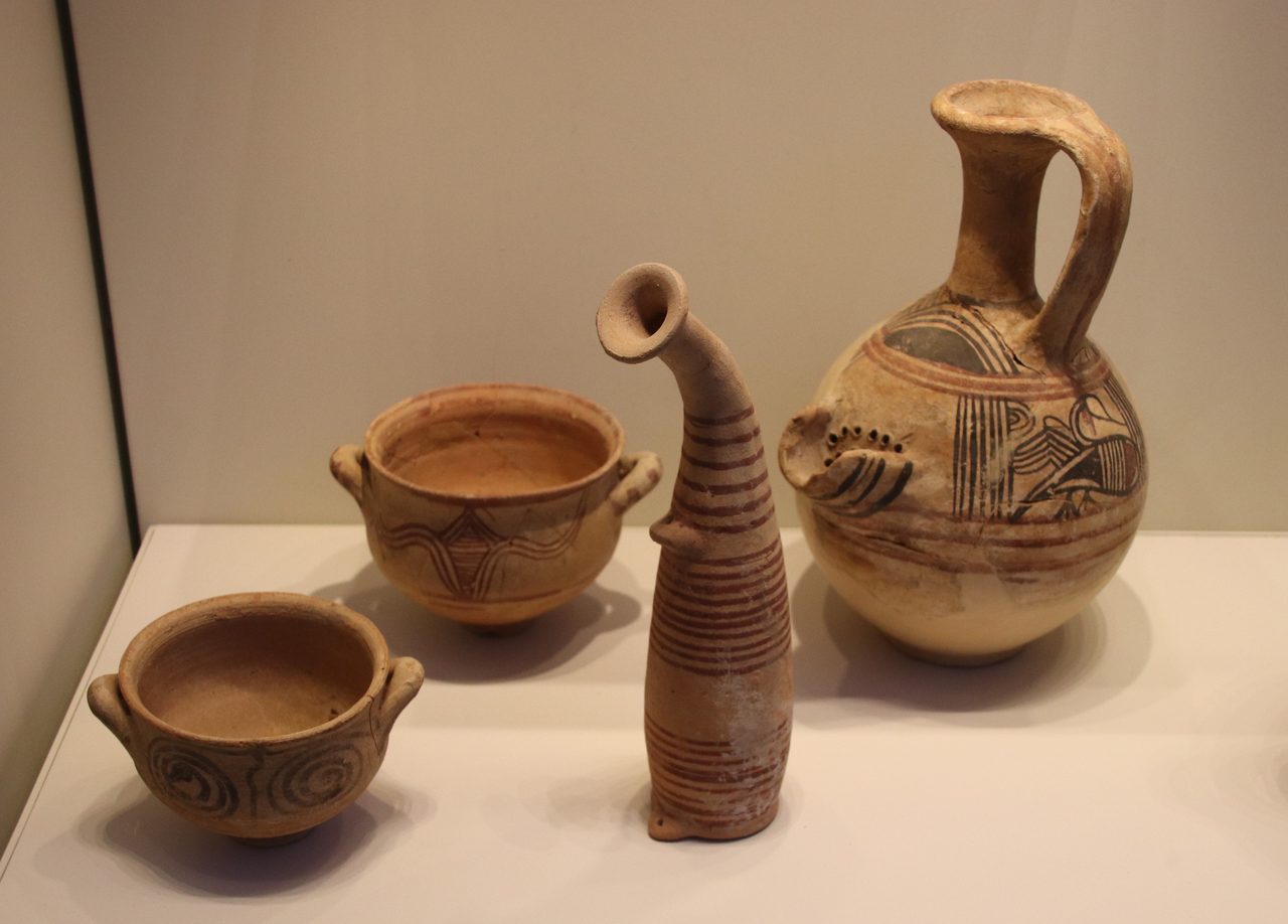 Four painted clay vessels on a white museum shelf: at the front a small two handled bowl with two large dark spirals, behind it a larger two handled bowl with a hatched chevron, in the middle a tall curved horn shaped bottle ringed with many horizontal bands, and at the right a round bellied jug with a tall neck, a strap handle, a spout pierced with small holes and a long necked bird painted on the body