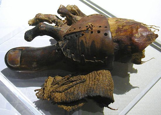 A mummified human foot with a dark wooden prosthetic big toe attached by a stitched leather sheath, resting beside a fringed linen wrapping