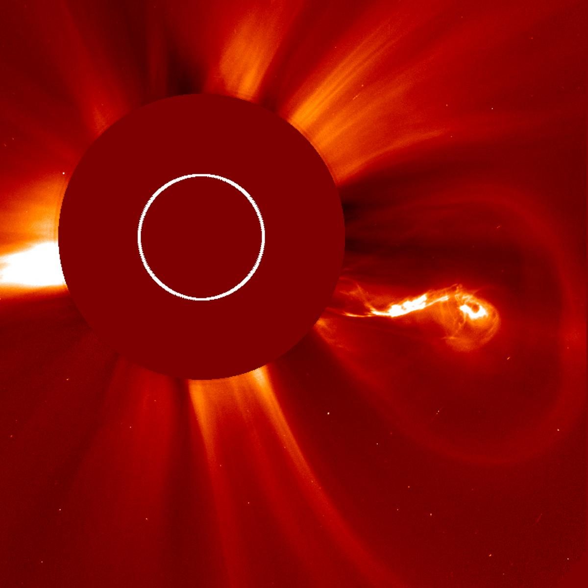 A square coronagraph frame rendered in reds and oranges: a large opaque dark disc left of centre with a thin white circle drawn inside it, streamers of light combing outward all round, a blaze of white spilling from behind the disc on the left, and a bright knotted cloud curled at its outer end erupting to the right.