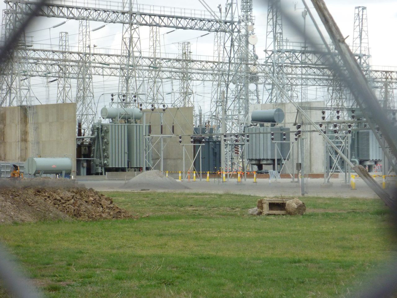 Three large grey transformers standing in separate concrete walled bays inside a transmission substation, ribbed cooling fins down their flanks and a horizontal cylindrical drum along the top of each, under a dense lattice of steel gantries and insulator strings, with mown grass and a spoil heap in the foreground.