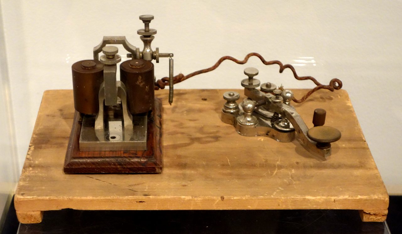Two brass and nickel telegraph instruments standing on a plain pale wooden board and joined by a rusted, wavy twisted wire: at the left a sounder with two upright cylindrical coils on a metal base, at the right a key with a long lever, knurled adjusting screws and a large round dark knob.