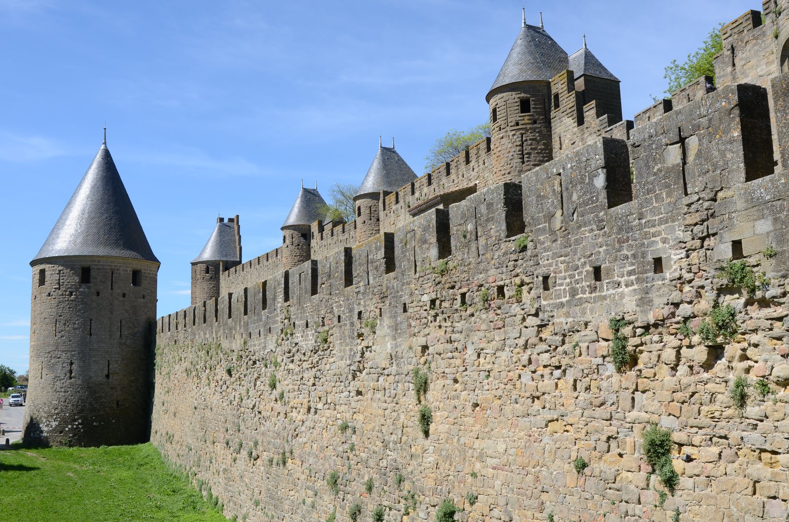 A long stretch of medieval curtain wall receding to the right under a bright blue sky, with five or six round towers carrying conical slate roofs, crenellated parapets and arrow slits. Lower courses are rougher stone with plants growing from the joints. A grassy ditch runs along the foot of the wall, and a white van and two small figures stand at the far left.