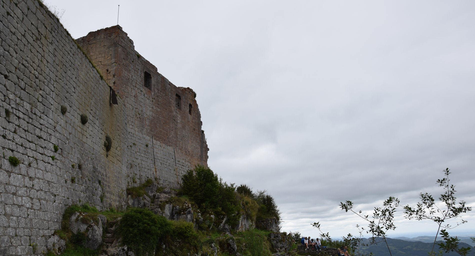 The curtain wall of a hilltop fortress rising steeply at the left in coursed pale stone, with a roofless keep above carrying window openings and a lightning rod. The ground falls away sharply to the right into a deep valley with distant ranges under a flat overcast sky. A worn footpath with rough stone steps climbs beside the wall, and five or six visitors sit on the rocks at lower right.