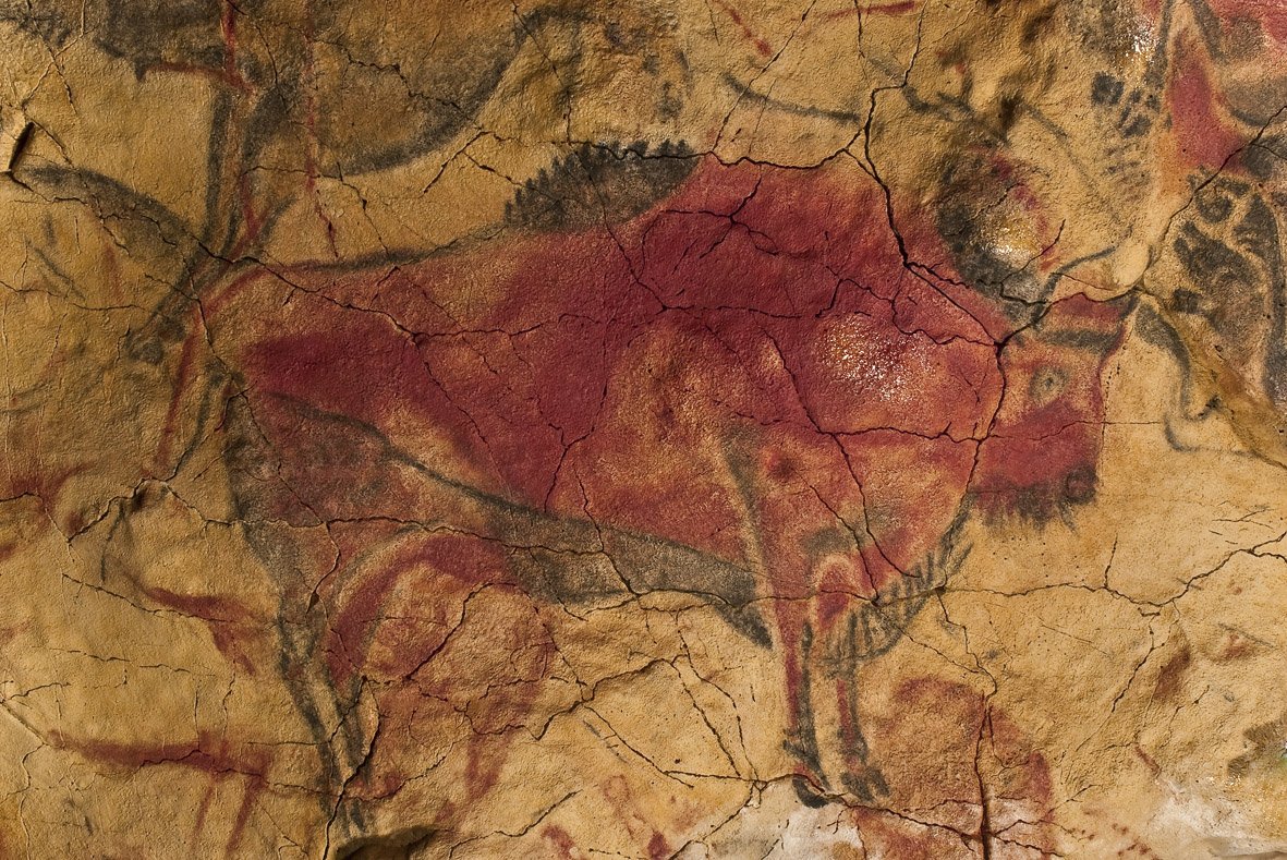 A large bison painted on a cracked tan cave ceiling, its body filled with deep red ochre and its head, horns, hump, and legs outlined in black, the natural swell of the rock giving the figure volume