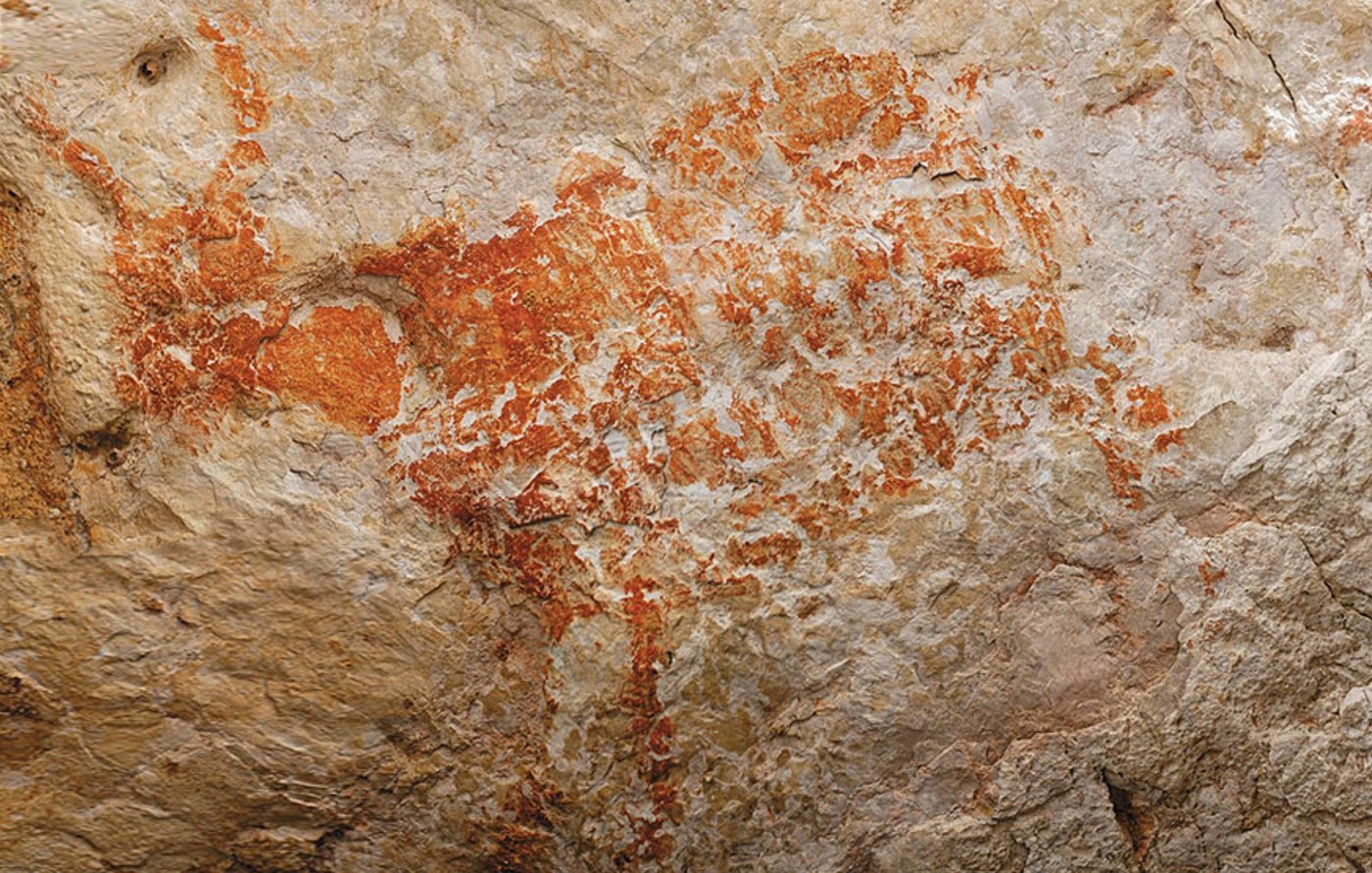 A red-orange painted figure of a heavy-bodied wild ox on pale, weathered limestone, its outline and body filled with ochre pigment, patchy with age