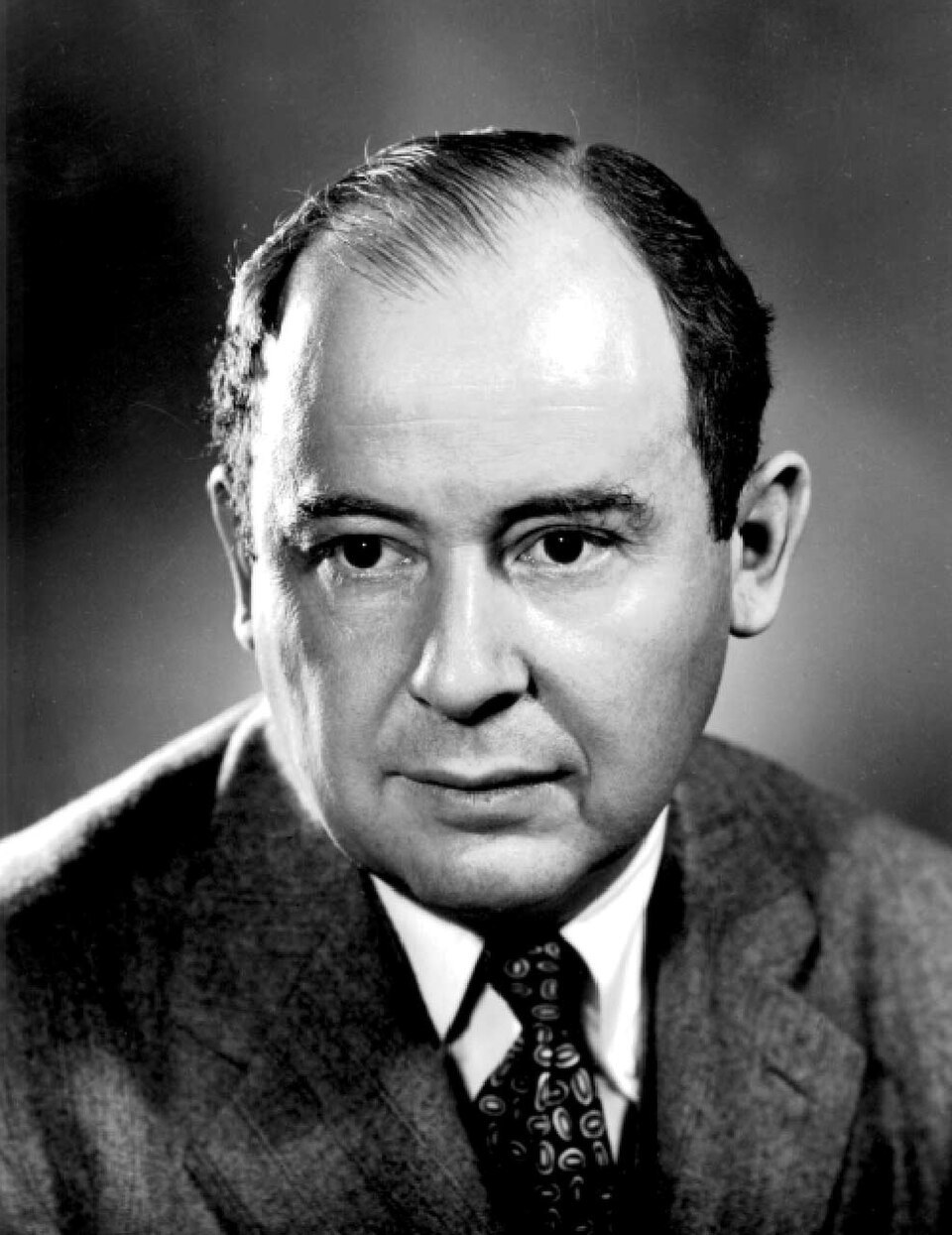 A black-and-white studio portrait photograph of the mathematician John von Neumann in a suit and patterned tie, with receding dark hair, against a grey background