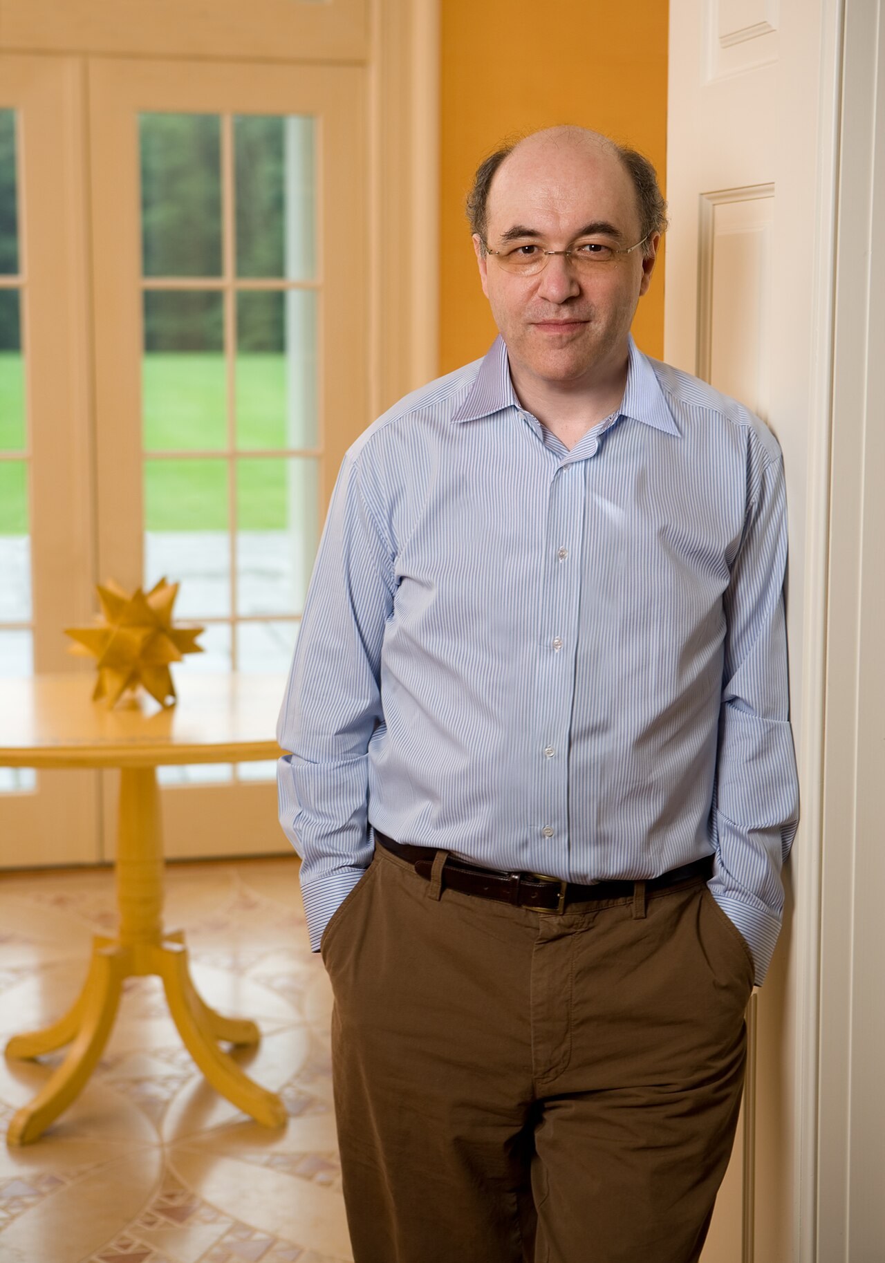 A photograph of Stephen Wolfram, balding and wearing glasses and a blue striped shirt, standing with hands in his pockets and leaning against a doorframe in a warmly lit room, a small star-shaped sculpture on a table behind him