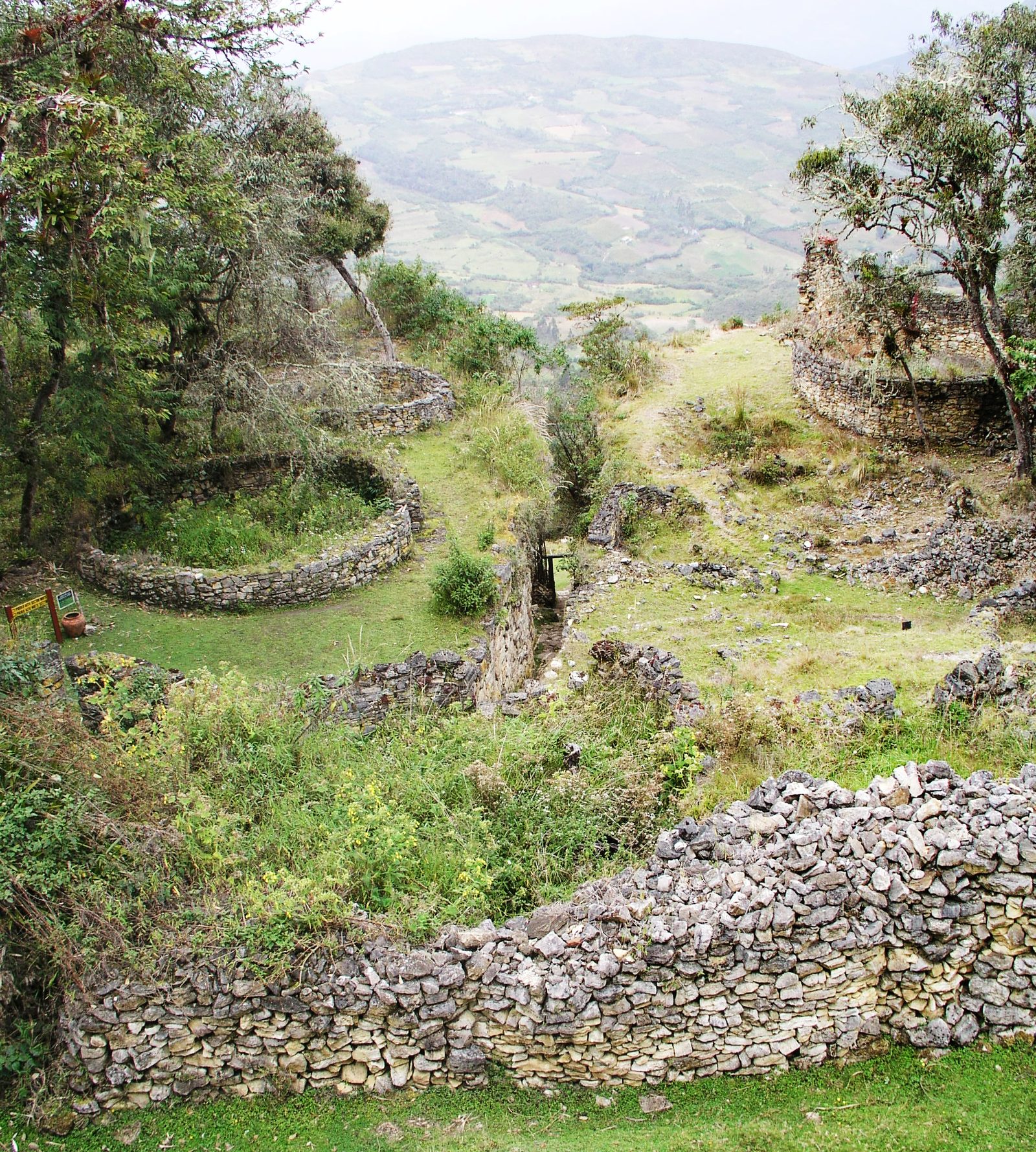 An overview of a ruined settlement on a terraced hillside. Several complete rings of dry-stone walling stand in green grass, the foundations of round buildings, one behind another. A taller round tower with intact walls sits at the upper right among trees. A long stone retaining wall crosses the foreground. A cultivated valley and further hills recede into haze beyond. A small interpretive sign and a ceramic pot stand at the left edge.
