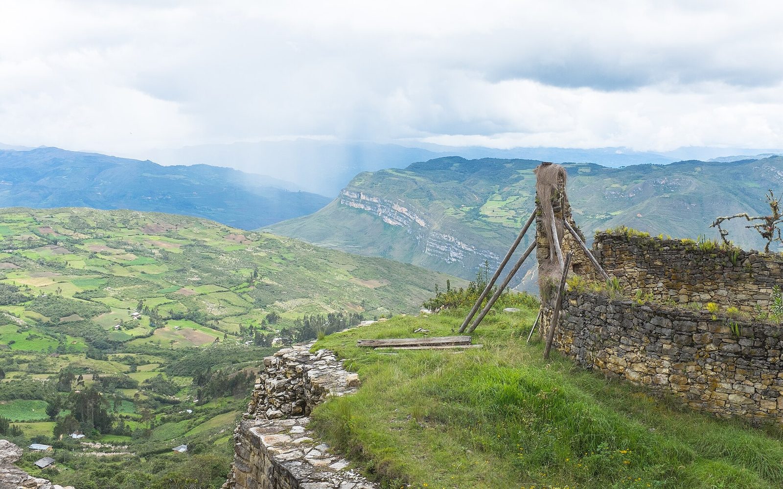A wide view out from a hilltop site. A patchwork of cultivated green fields fills a valley far below with scattered farm buildings. Rain falls in visible shafts from low cloud over ridges in the middle distance, and pale limestone escarpments band the hillsides. At the right, the top of a stone wall, and standing on it a round structure with a conical thatched roof held upright by long timber props.