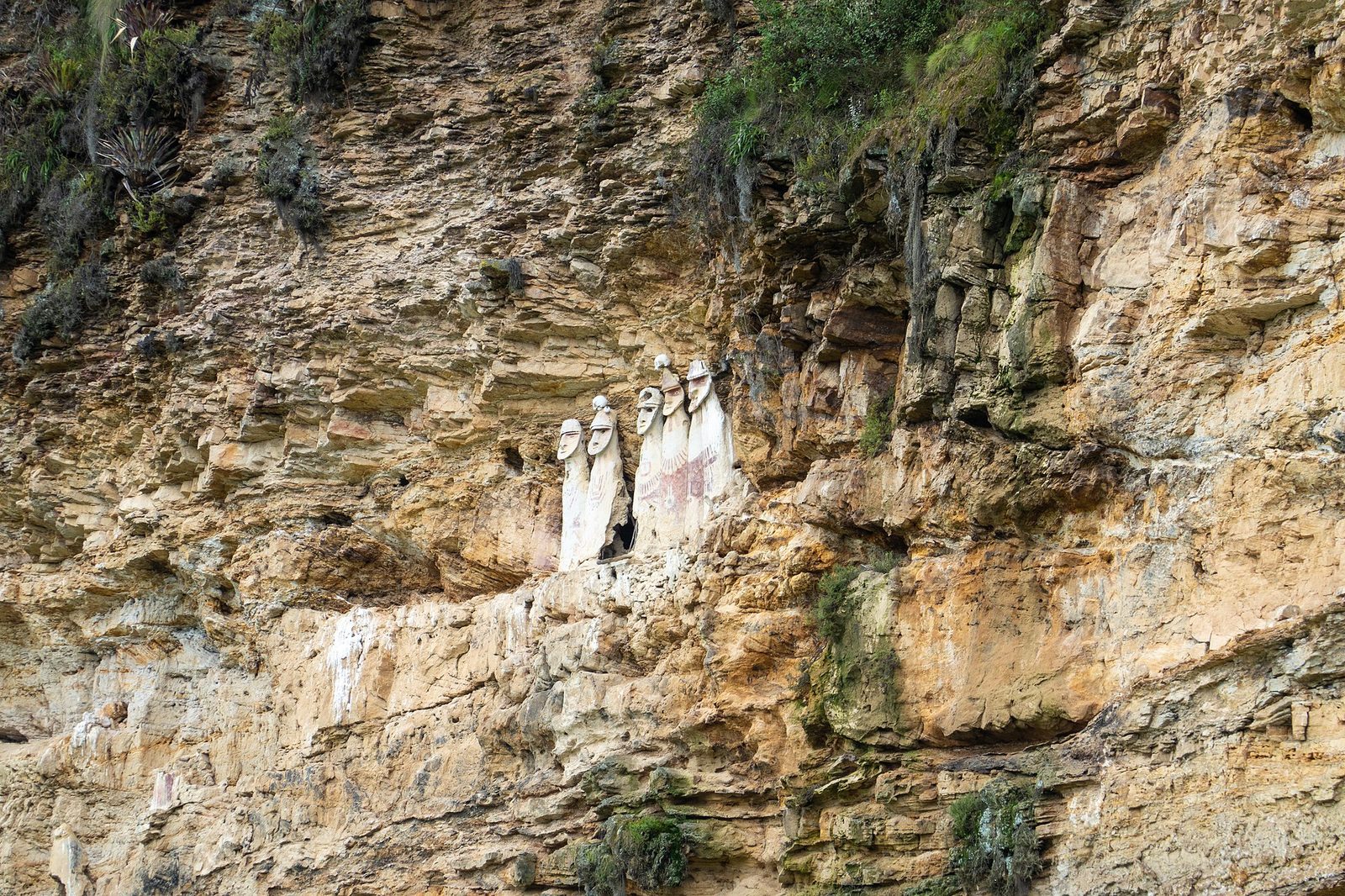 A vast tan limestone cliff face, layered and pocketed, filling the whole frame. On a narrow ledge left of centre stand six pale elongated human-shaped figures, shoulder to shoulder, with modelled faces and flat helmet-like tops, some carrying red painted decoration on their bodies. One has a dark opening in its side. Scrubby vegetation clings to the rock above and below them.