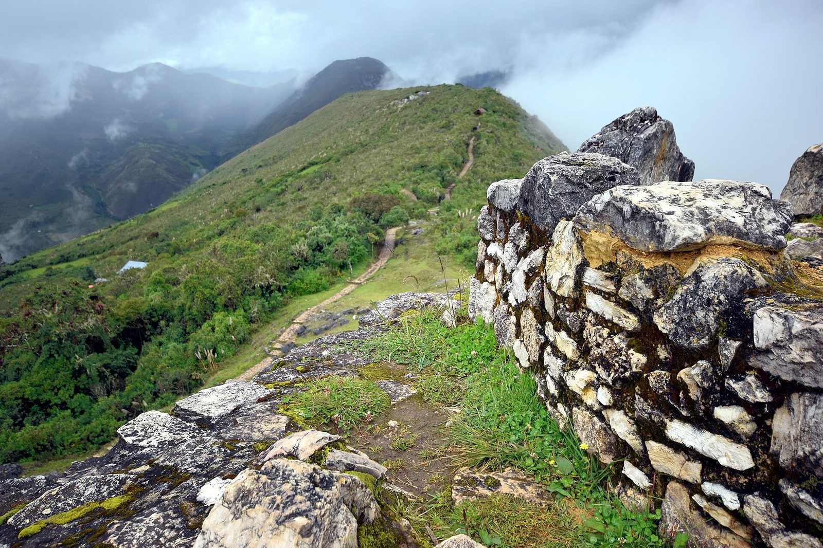 A view from on top of a massive stone wall. Coursed pale limestone masonry fills the right of the frame, with a mossy stone parapet running away to the lower left. Beyond it a steep green ridge falls away, a footpath switchbacking down it, a single small blue-roofed building in the middle distance, and cloud pouring over dark mountains along the whole horizon.
