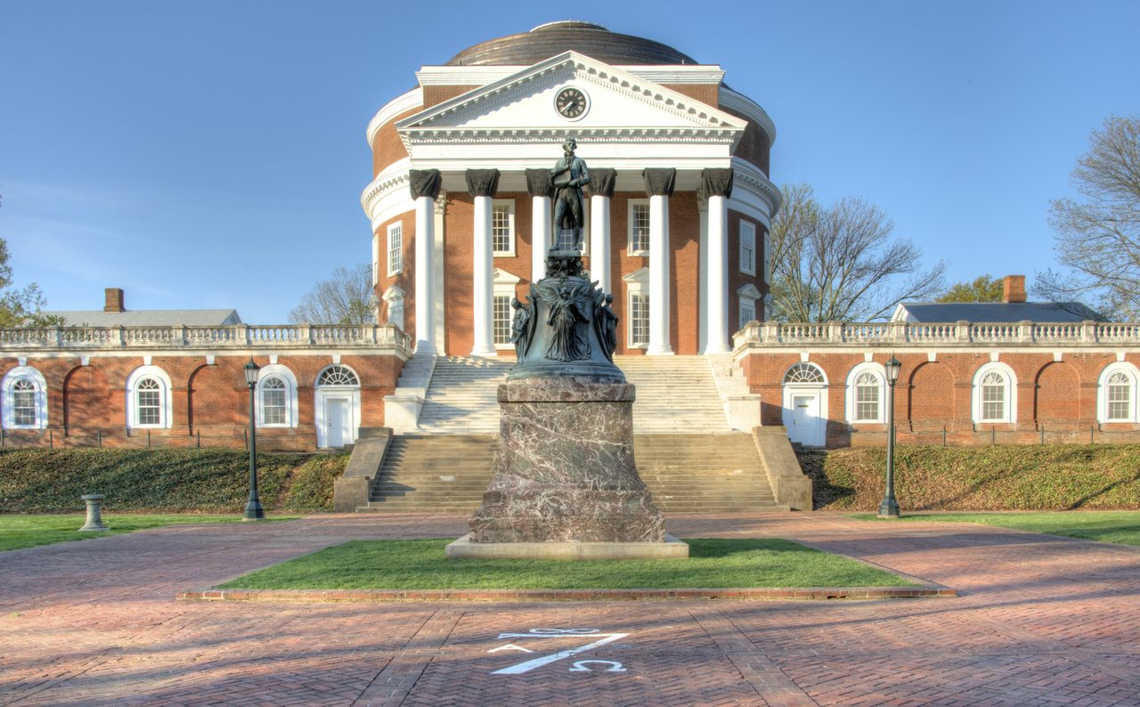 The domed brick Rotunda at the University of Virginia behind a white-columned portico, a bronze statue on a tall stone pedestal standing in front of its steps, a dark cover fitted over the top of each column, and low brick colonnades running away to either side