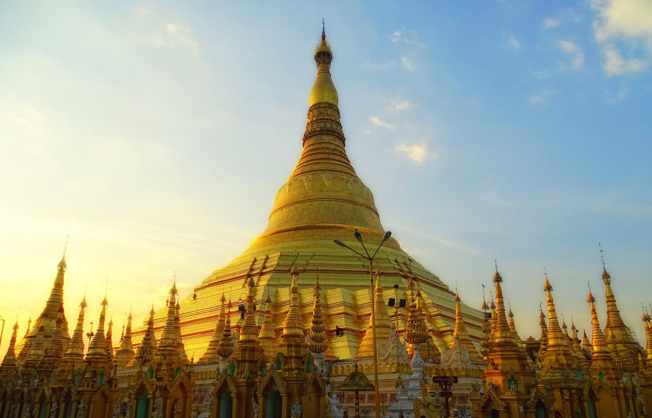 The gilded Shwedagon Pagoda stupa rising in low golden sunlight above a dense ring of smaller golden spires, a tall lamp standard with three lamp heads crossing the gold in front of it