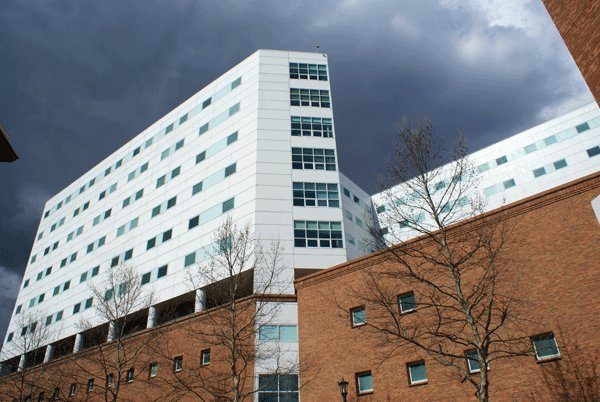 A white panelled hospital tower photographed steeply from below against a dark overcast sky, with a red brick building and bare trees across the foreground