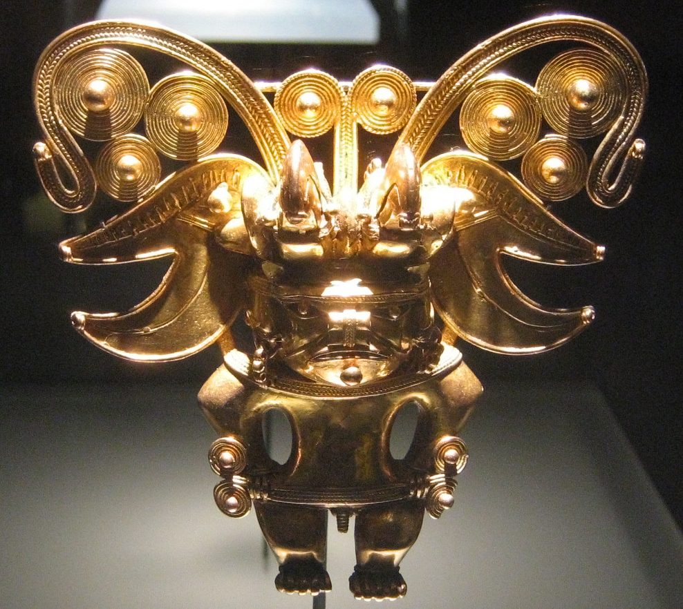 A cast gold pendant glowing against a dark museum case. A squat frontal figure wears a wide crested headdress built from coiled spiral discs and outward-curving crescent plates, with two upright projections above the face. Spiral ornaments hang at the ears and at the hips, a belt crosses the waist, and small feet show at the bottom. Fine coiled wire work is visible across the whole piece.