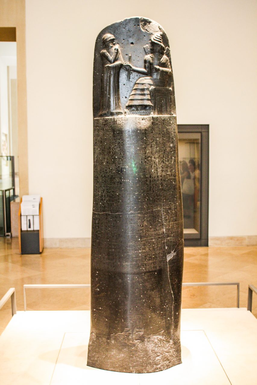 A tall black stone stele covered in cuneiform inscription, with a carved relief scene at the top