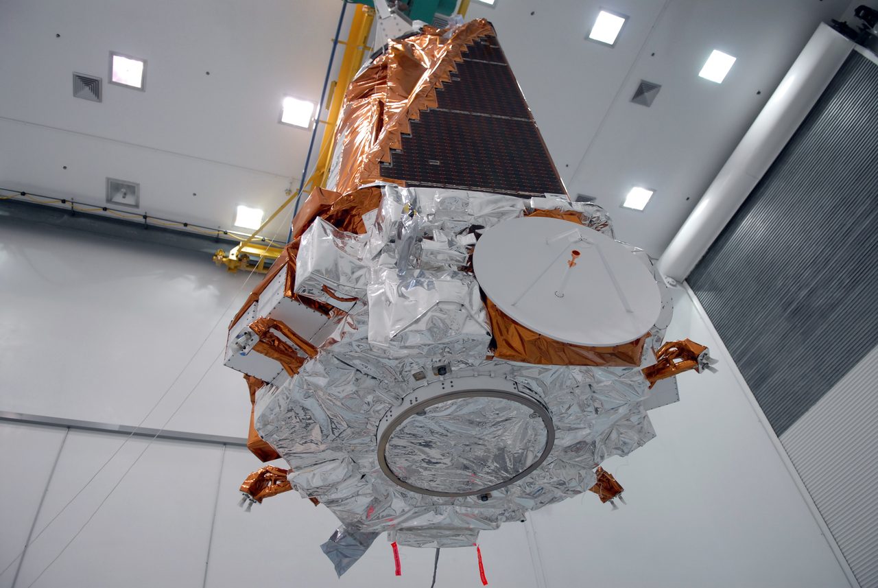 The Kepler spacecraft suspended from handling equipment inside an empty white processing bay, its solar array, dish and launch-vehicle mating ring visible