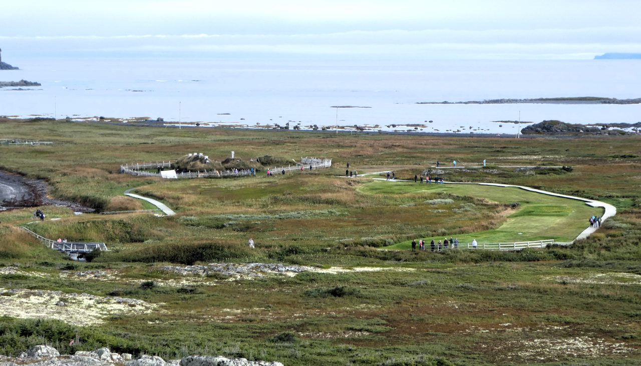 A coastal meadow at L'Anse aux Meadows in Newfoundland, with low grassy ground in the foreground and turf-roofed buildings in the distance under an open sky