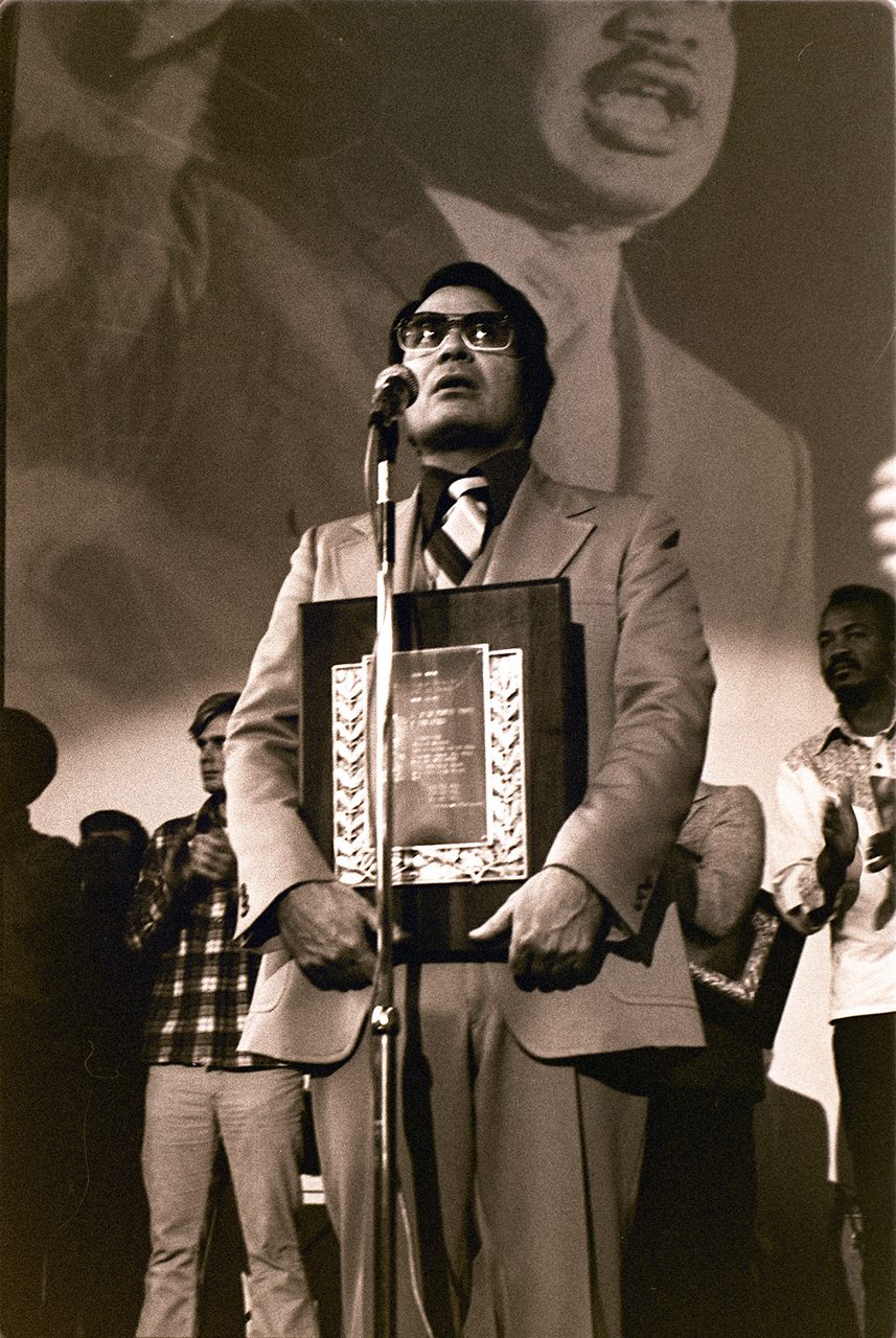 A sepia toned photograph in portrait format: a man in a light coloured suit, dark tinted glasses and a dark patterned tie stands at a microphone on a stand, holding a large wooden plaque against his chest whose engraved inner plate is framed by a leaf wreath border; behind him a very large photographic backdrop shows the head and shoulders of a moustached man in a suit and tie, and other people stand at the edges of the frame, partly in shadow.