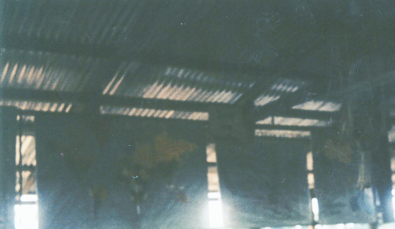 A dark, grainy colour photograph with a heavy blue green cast, looking into a large open sided shelter: corrugated roofing fills the top of the frame above a run of horizontal beams and rafters, upright posts drop away into deep shadow below them, and narrow bands of daylight show through the gaps at ground level along the bottom edge; the film is scratched and fogged and the lower half of the frame holds no legible detail.