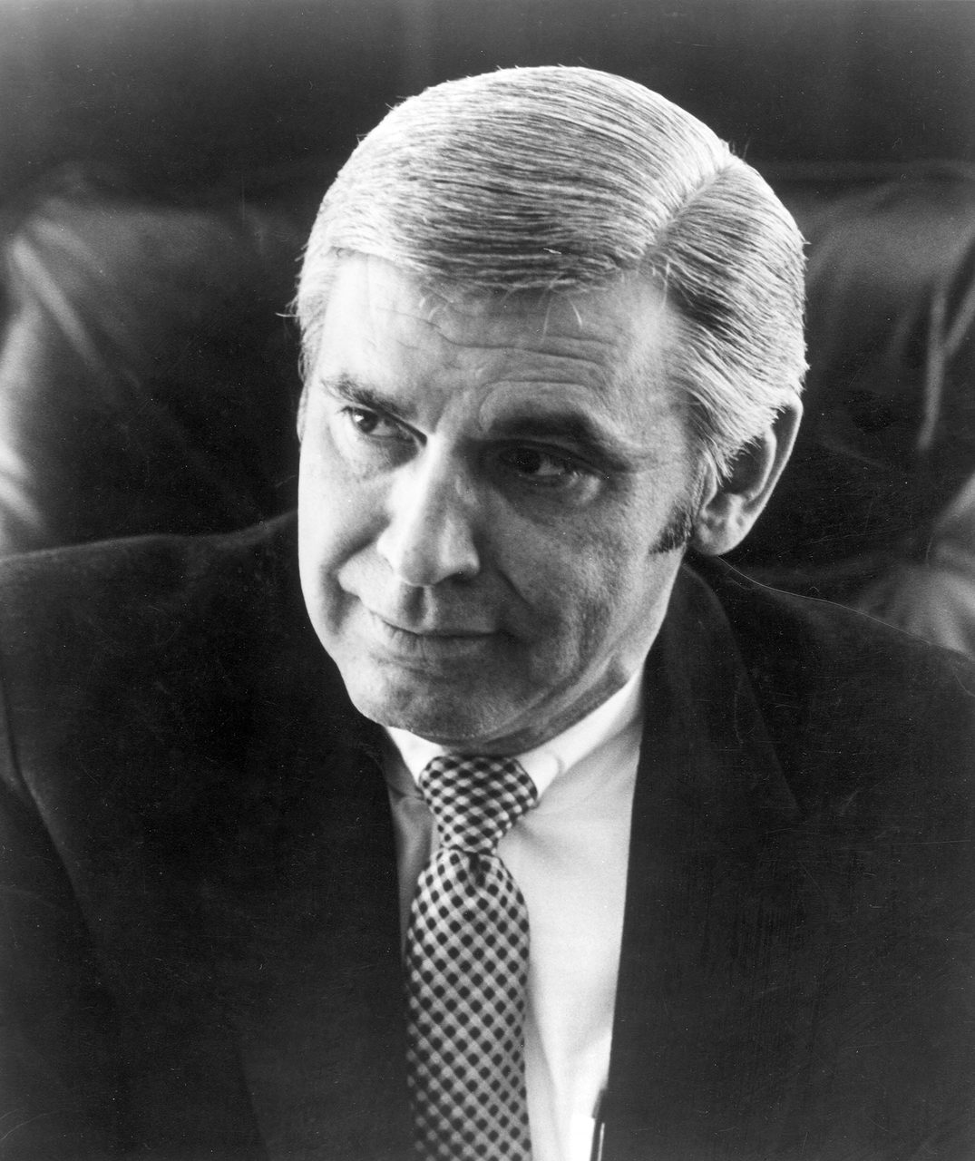 A black and white studio portrait of a man in late middle age, shown from the chest up against the dark padded back of a leather chair: swept back light coloured hair, a dark suit jacket, a white shirt and a dark tie with a small check pattern, his head turned slightly to one side.