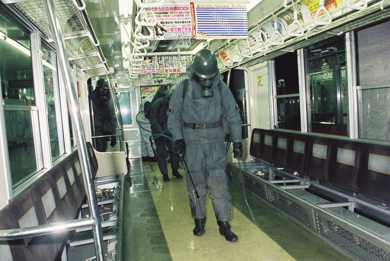 A colour photograph inside a subway car with no passengers in it: a figure in a full grey protective suit, gloves, boots, a dark helmet and a gas mask walks down the centre aisle holding a spray wand on a hose, with liquid standing on the floor; more figures in the same equipment work further along the car; strap handles, hanging advertising cards and bench seats line both sides and the windows are dark.